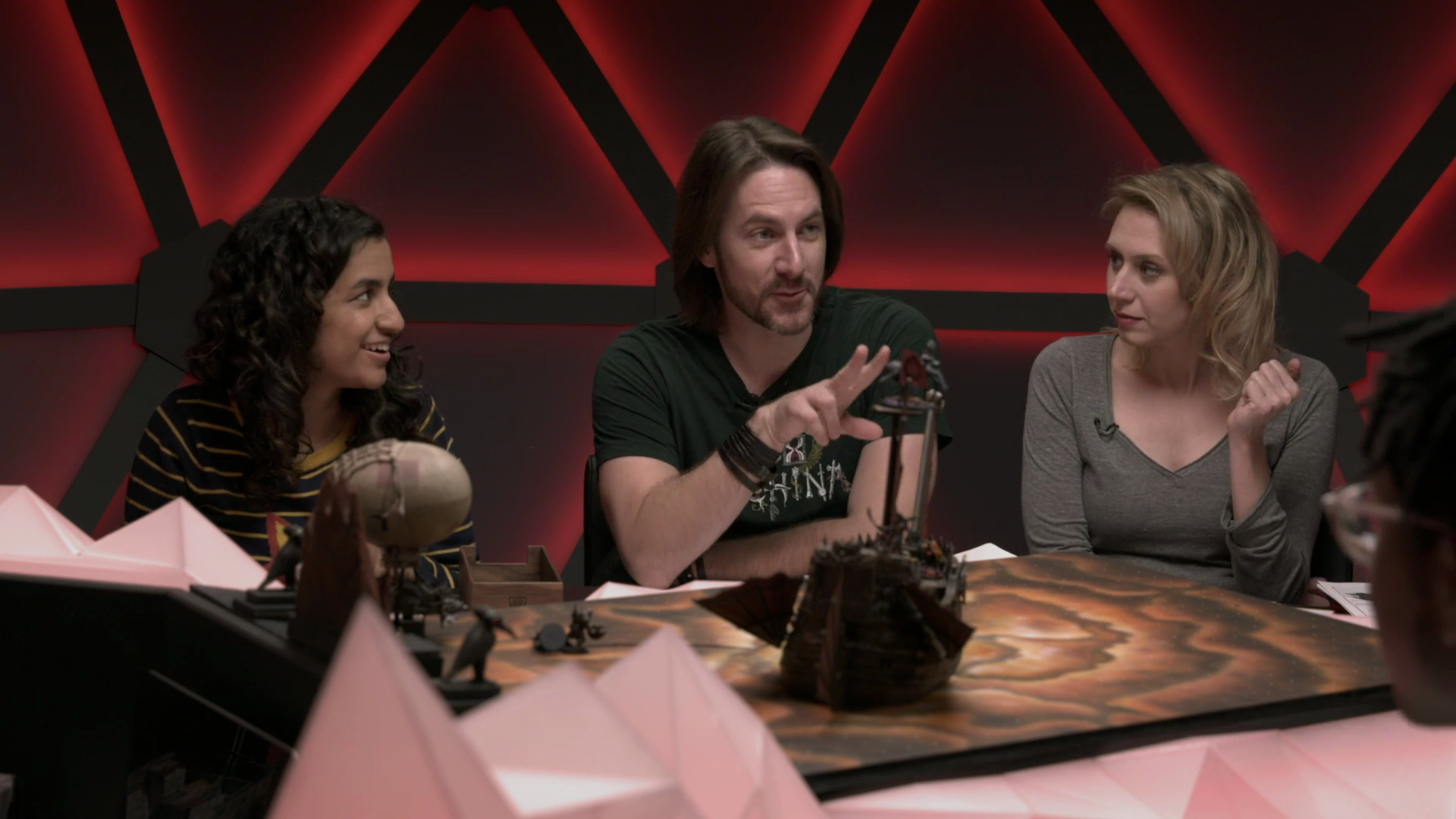 Three people sit around a table playing a tabletop role-playing game, with miniature figures and map in front of them. The person in the middle, with long brown hair and a beard, is gesturing with his hand, while the woman on the right with blonde ha