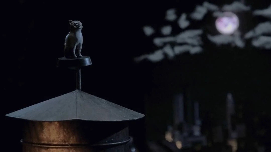 Close-up of a black cat figurine perched on top of a metal water tower at night, with blurred trees and moon in the background.