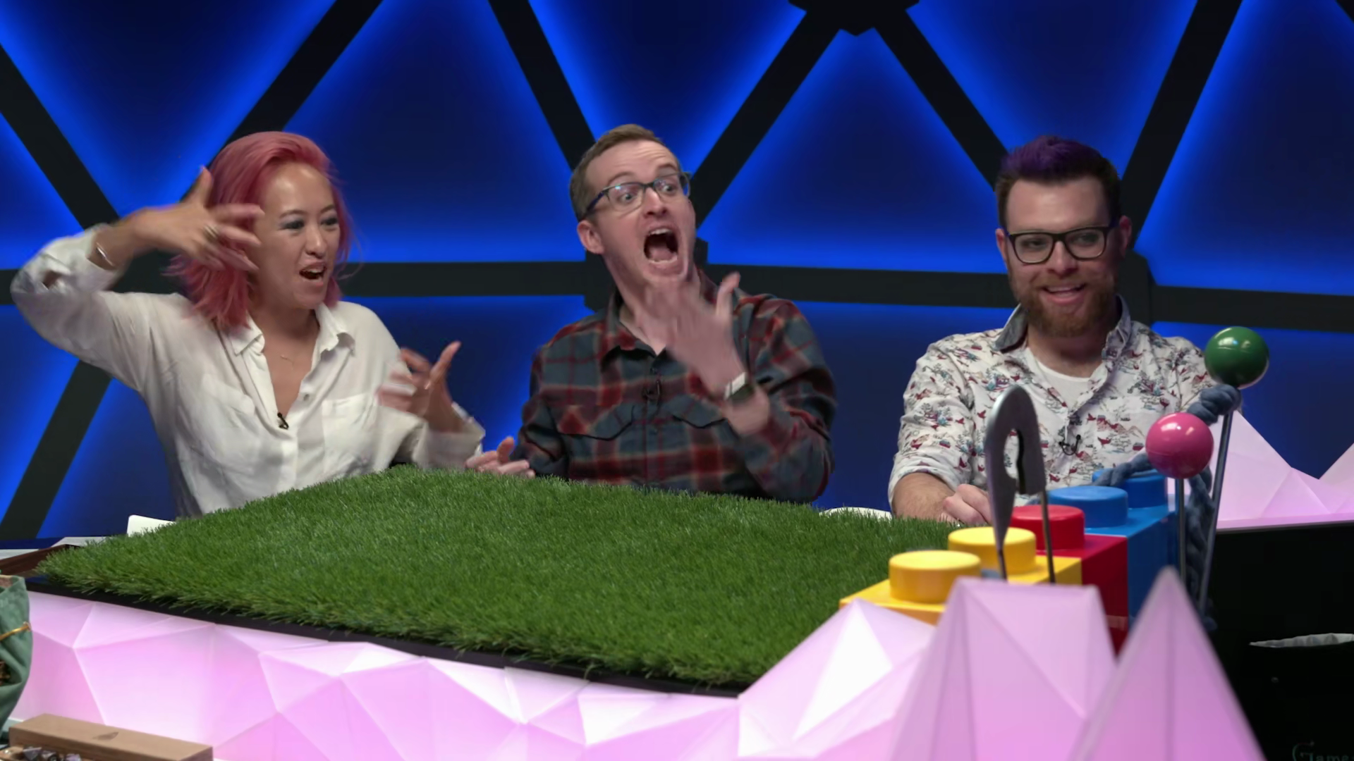 Three people engaged in a lively discussion at a game show set with colorful props, including a grassy table and geometric decorations, against a blue and black background.