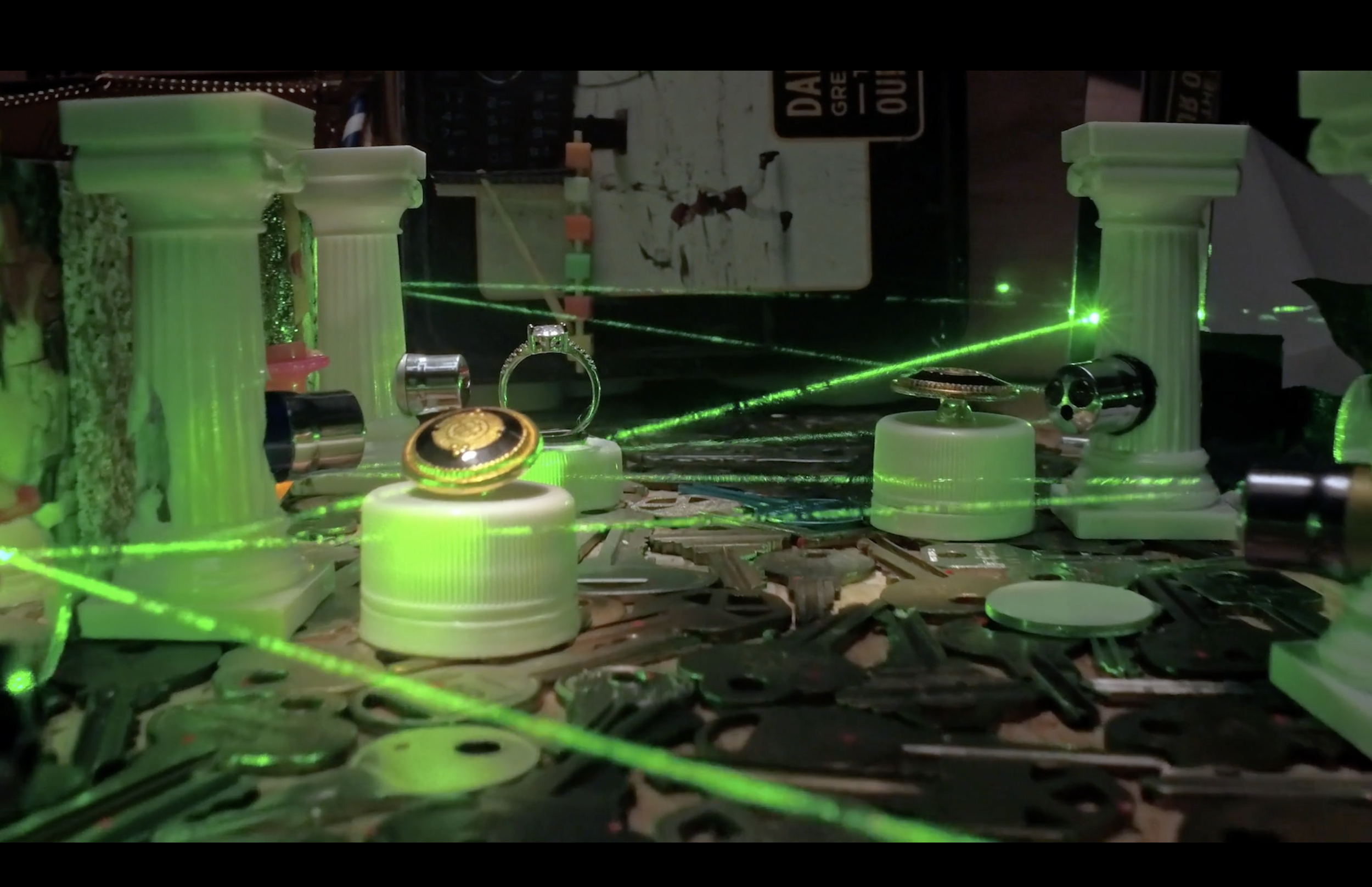 A miniature heist scene with green laser beams, white columns, and various metallic and plastic objects scattered around. A diamond ring is present on a platform.