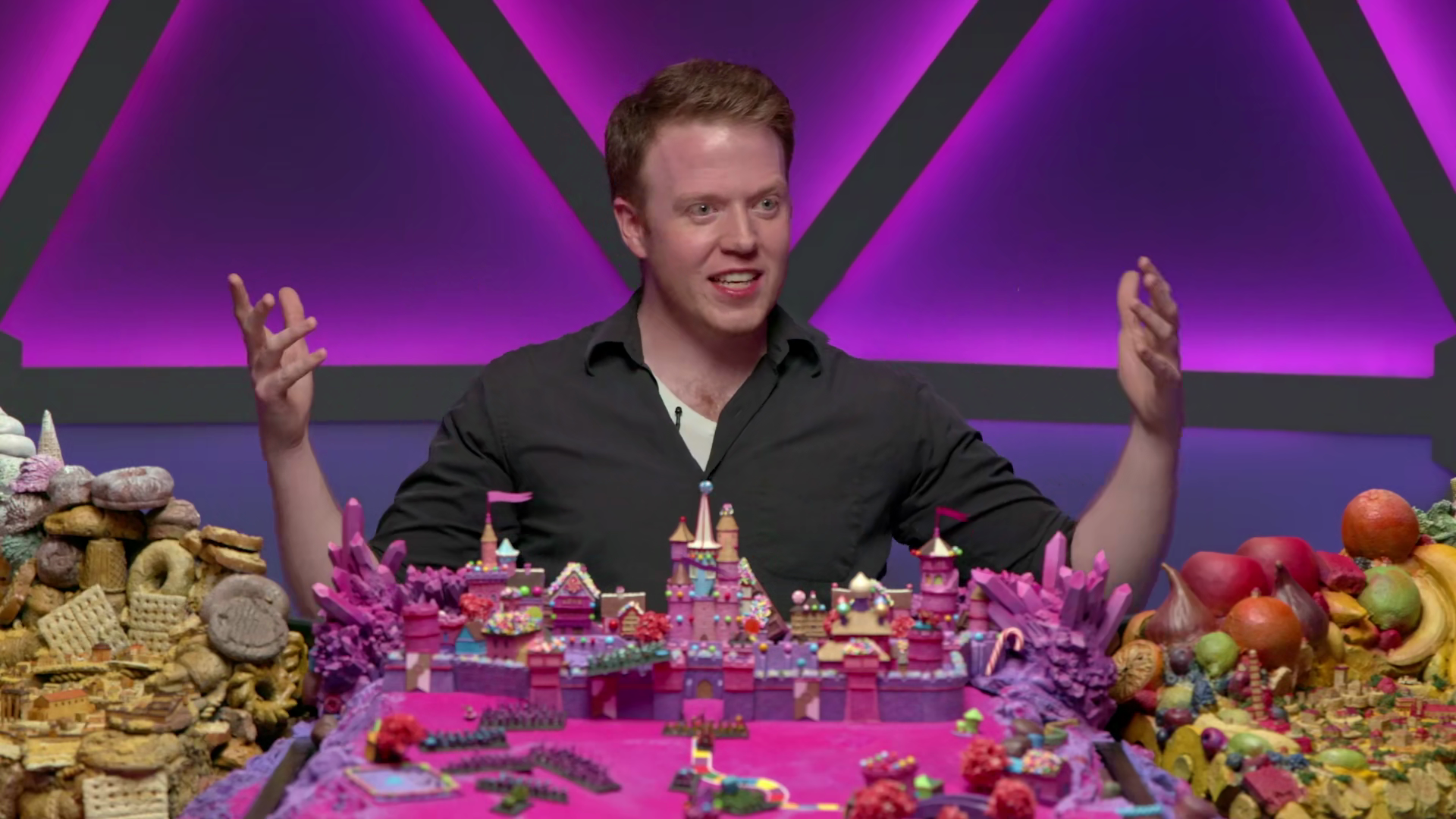 A man with red hair and a black shirt sitting at a colorful table filled with sweet treats, candies, and desserts, speaking and gesturing with his hands against a purple and black geometric background.