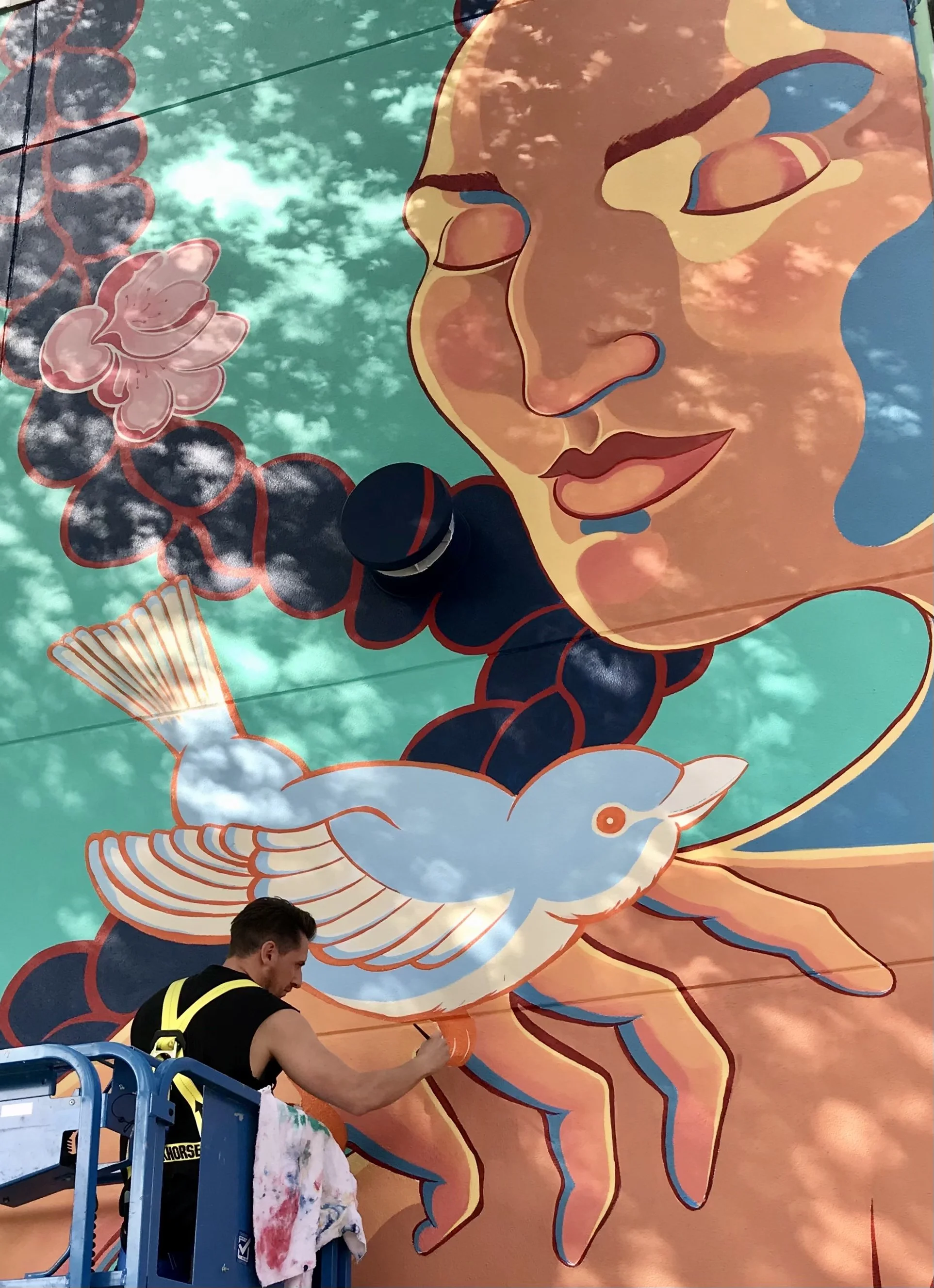 Mural artist, David Camisa, standing on a scissor lift while painting a colourful mural featuring a woman's face, a bird, and abstract floral designs on a large outdoor wall, with clouds reflected on the surface.