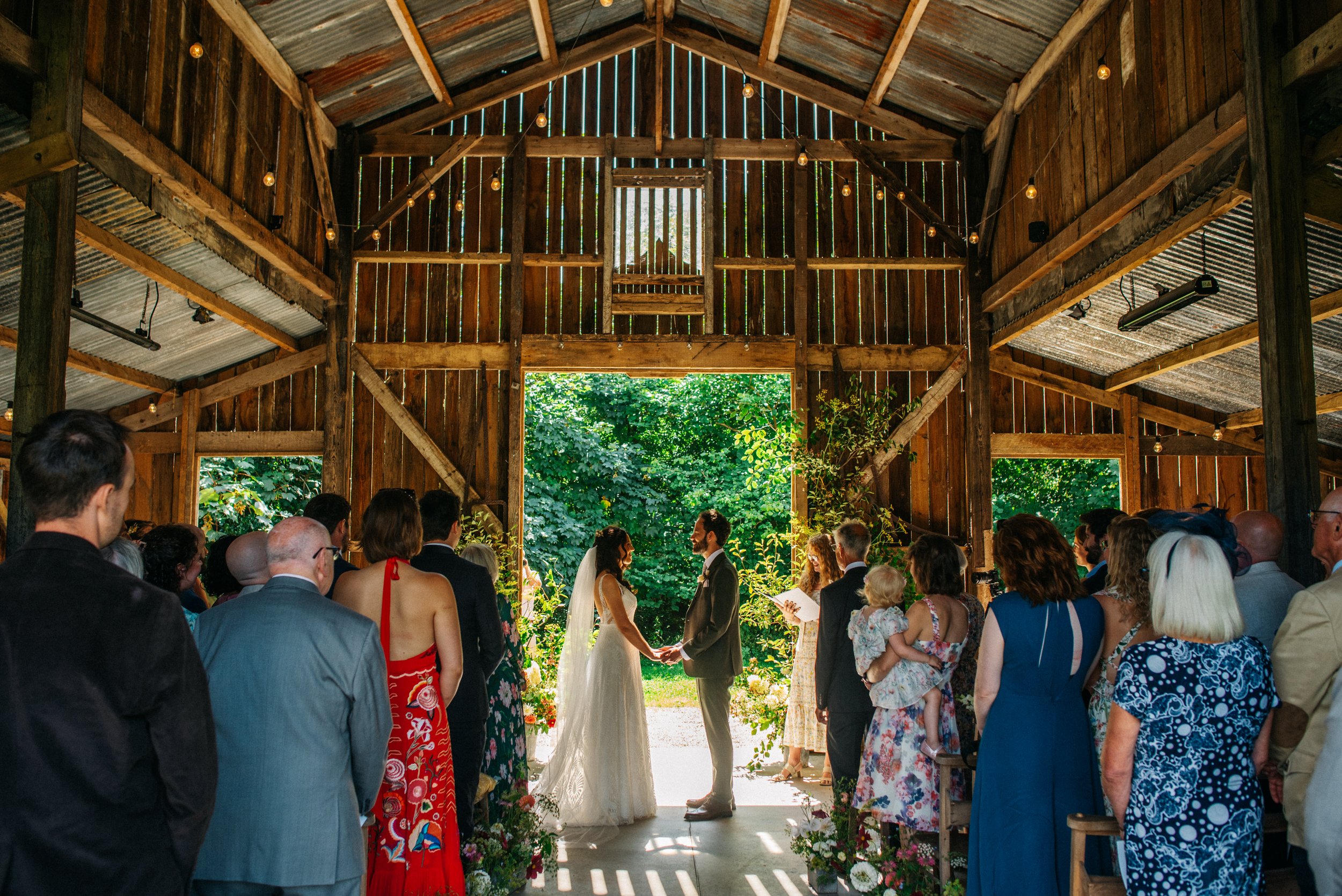 A wedding ceremony taking place inside a rustic barn with wooden walls and ceiling. The bride and groom are facing each other, holding hands, in front of the officiant, with guests standing on either side watching. Bright natural light is streaming i