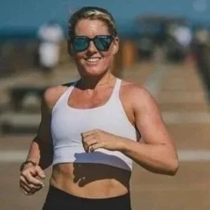 Woman running outdoors in athletic wear, wearing sunglasses, smiling, with a blurred outdoor background.