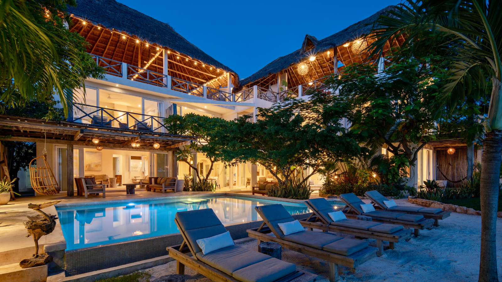 Luxury beachside villa at night with lit pool, lounge chairs, trees, and thatched roof structures.