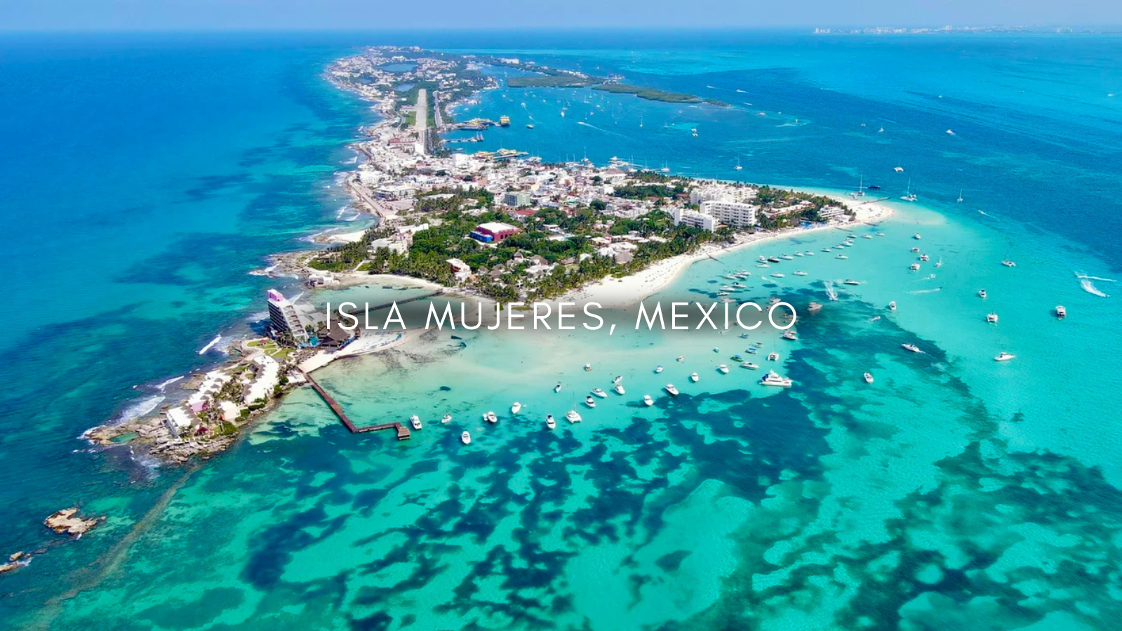 Aerial view of Isla Mujeres, Mexico, showing turquoise waters, boats, a sandy beach, and a resort on a small peninsula.