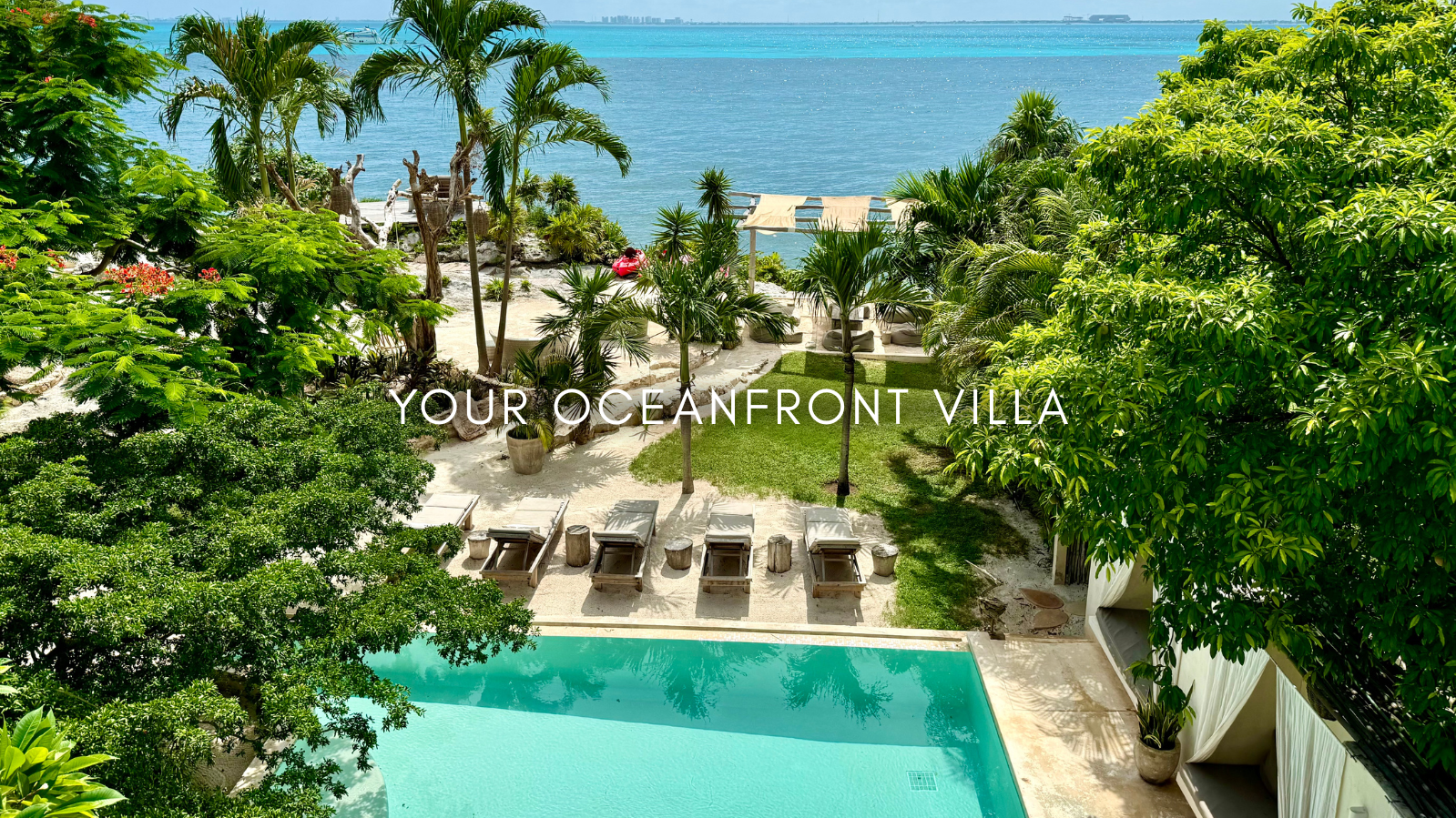 A lush oceanfront villa with a swimming pool, surrounded by tropical trees and plants, overlooking the ocean with a sandy beach and lounge chairs.