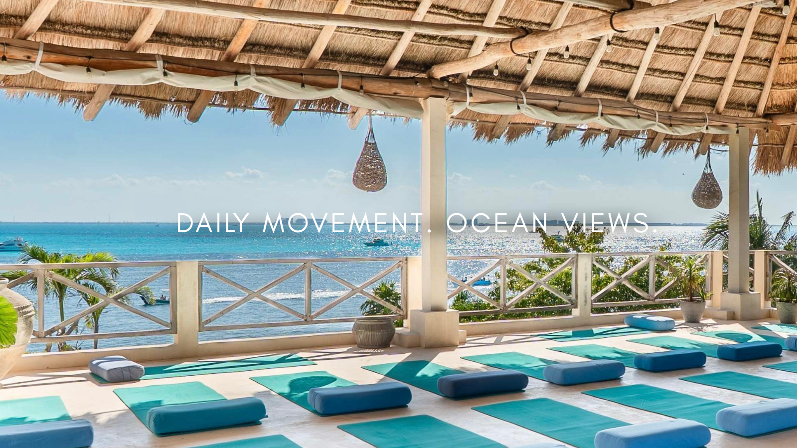 A beachside yoga deck with turquoise mats and cushions, overlooking the ocean with boats and palm trees, under a thatched roof with hanging wicker lamps.