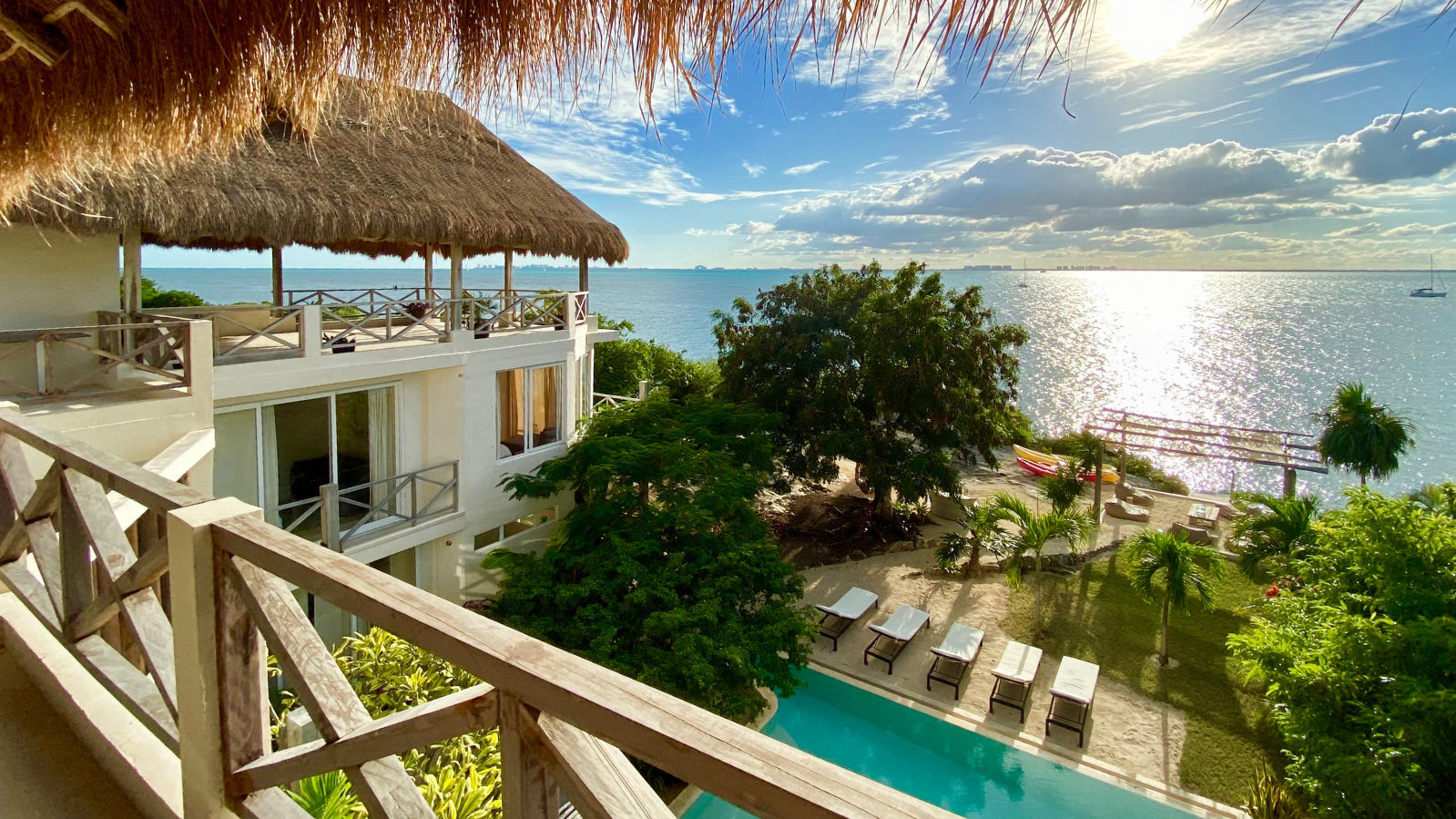 A tropical resort with a white building, a thatched roof, a pool, palm trees, and a view of the ocean with sailboats, under a partly cloudy sky.
