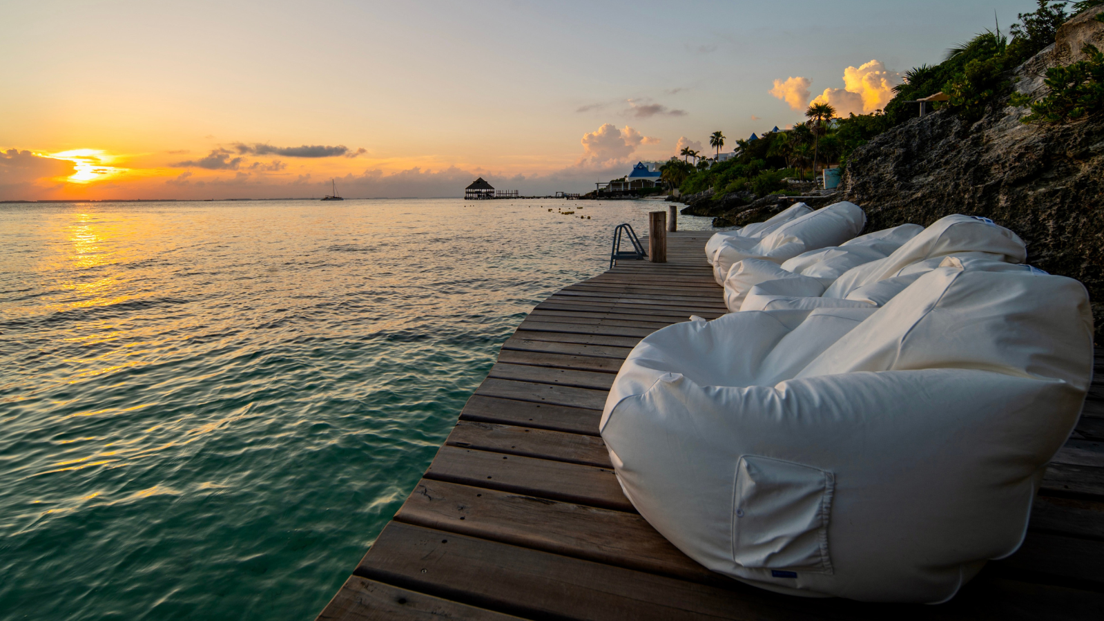 Beachside wooden dock with white bean bag chairs, rocky hillside with palm trees, and sunset over calm water with boats in the distance.