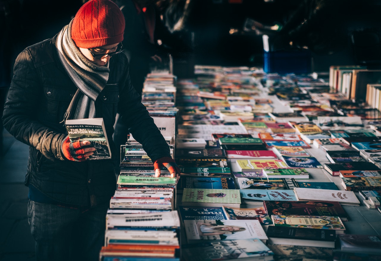 Photo: A man in a hat and scarf holding a book in one hand while browsing for other books on a table. 