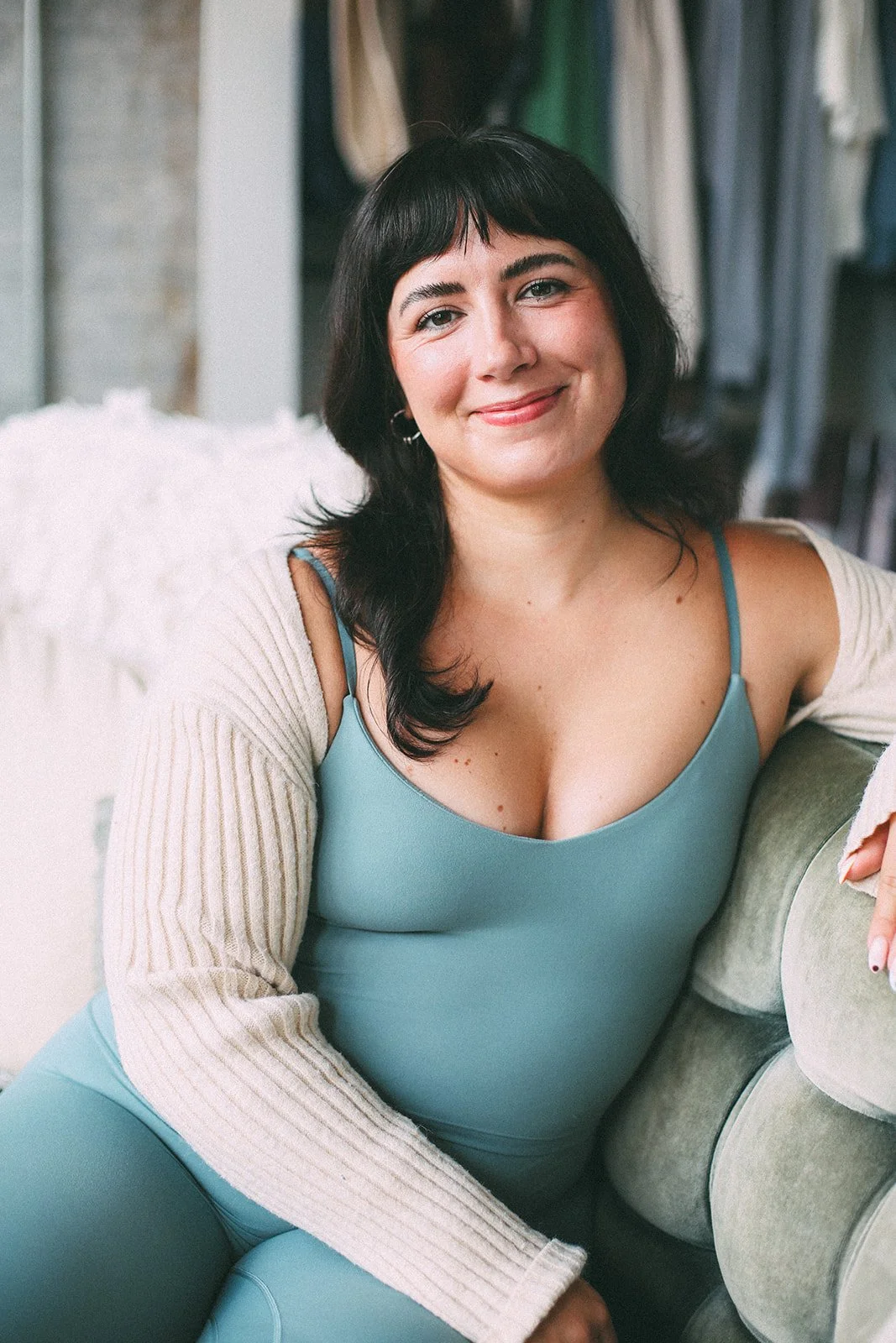 A woman with dark hair and bangs smiling, wearing a blue tank top and a beige cardigan, sitting on a green couch.