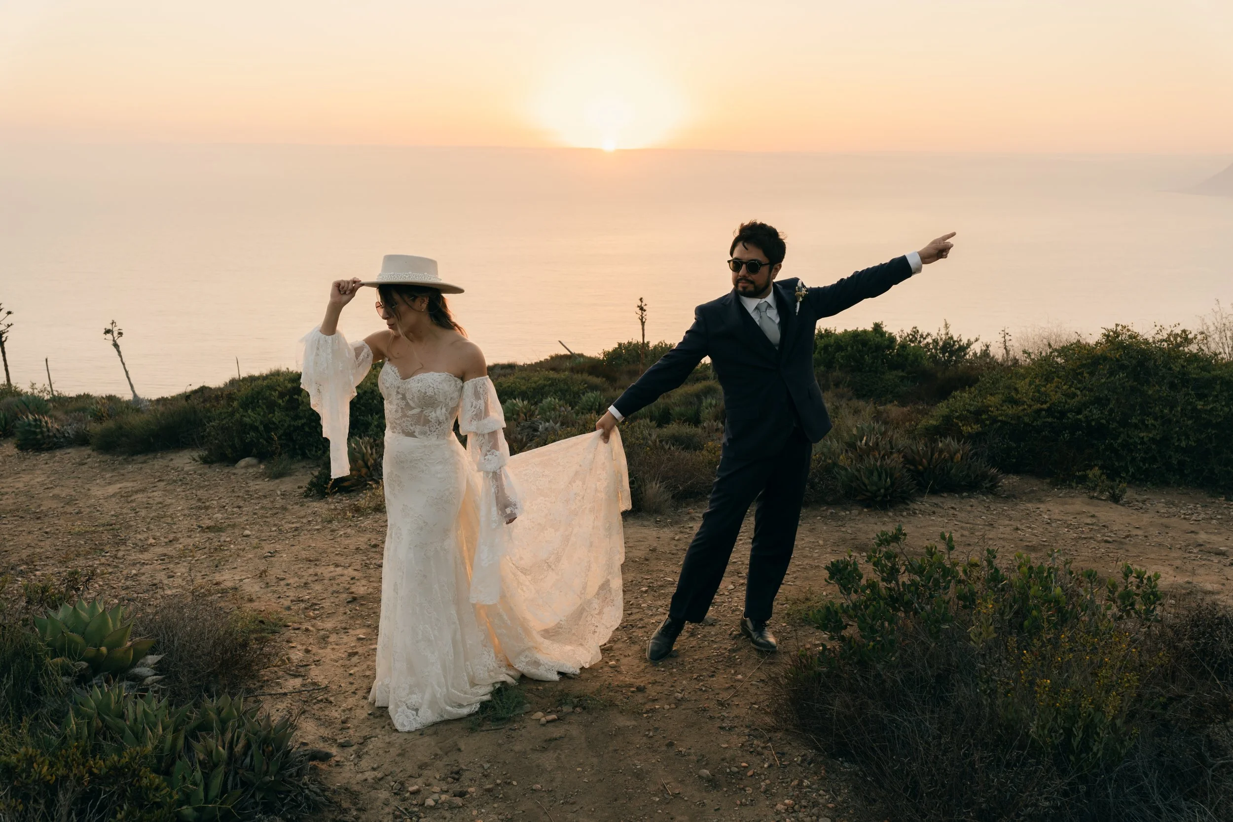 Pareja de recién casados en un paisaje al atardecer, con el océano y un horizonte en el fondo. La novia lleva vestido de encaje y sombrero, y el novio lleva traje oscuro y gafas de sol, en una pose romántica y divertida.