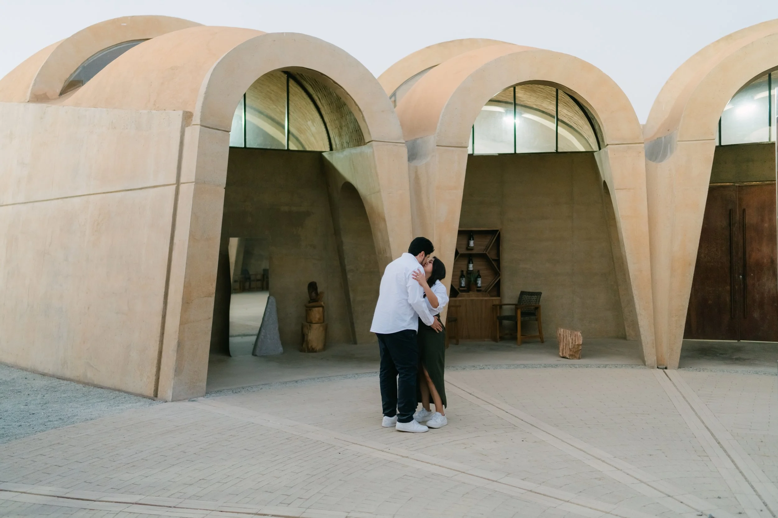Una pareja se besa frente a una estructura de arquitectura moderna de color beige con arcos y ventanas de forma semicircular, en un ambiente al aire libre.