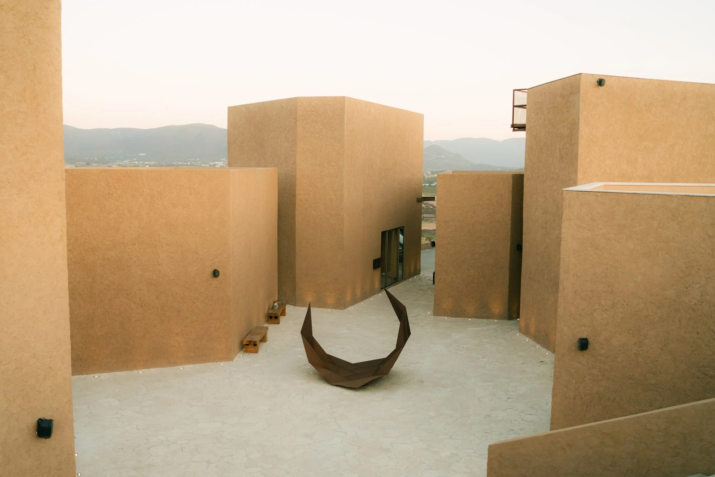 Vista de una estructura moderna con muros color terracota y una escultura de forma angular en el centro del patio, con montañas al fondo.