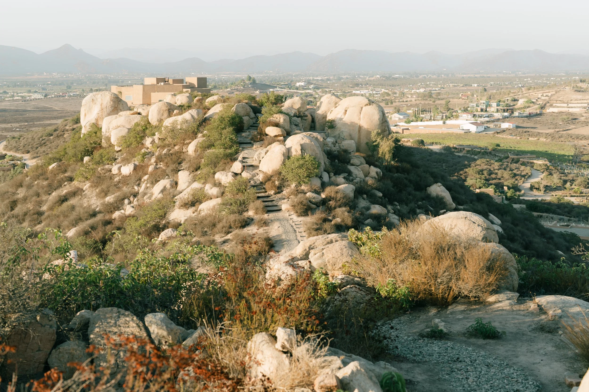 Camino de piedras en una colina con vegetación, rodeada de rocas grandes y arbustos, con una ciudad y montañas en el fondo.