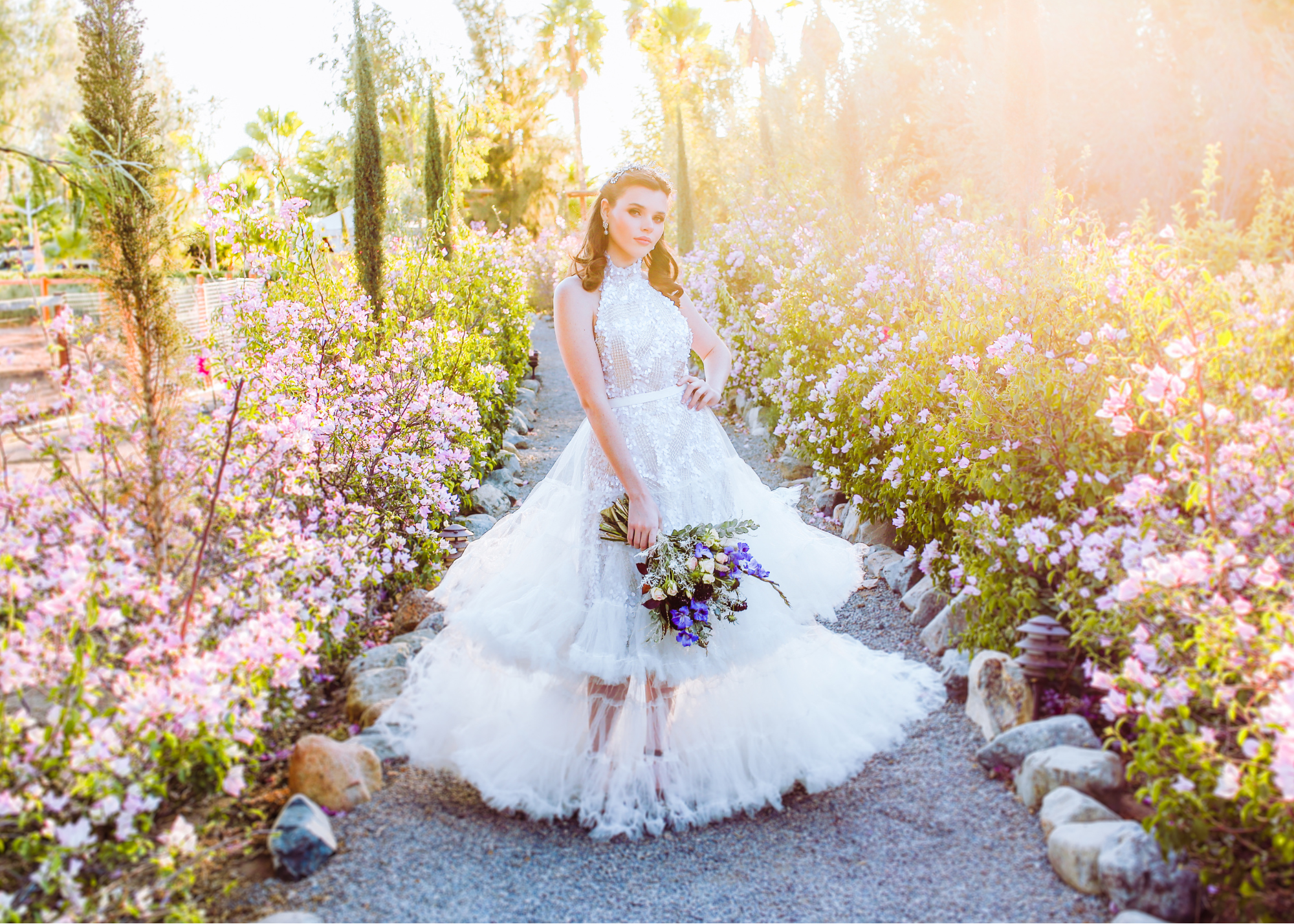 Mujer en vestido de novia en un sendero rodeado de flores rosas y verdes, con luz solar brillante