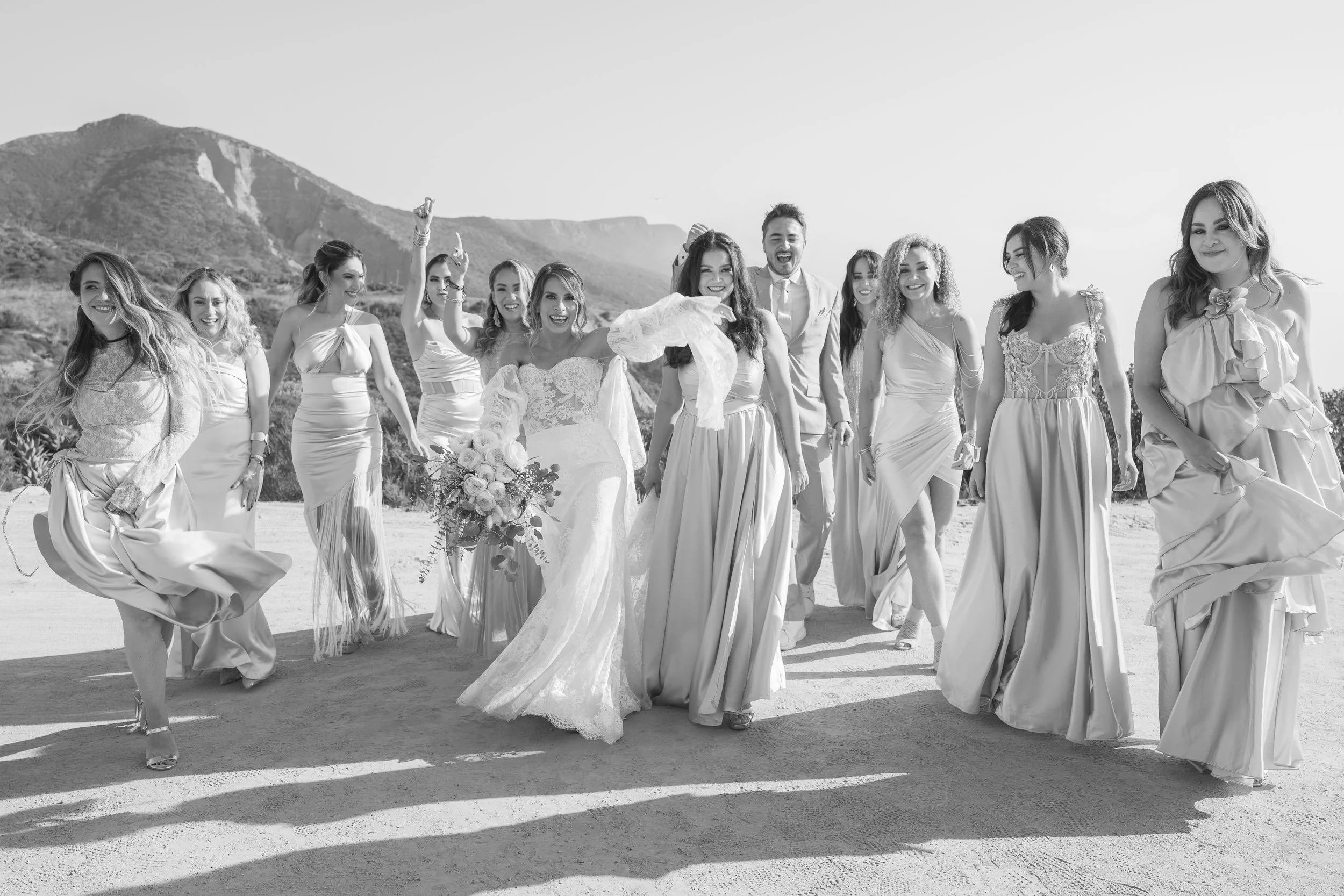 Grupo de personas en la playa, vestidos con ropa de boda, sonriendo y caminando hacia la cámara, con montañas al fondo.