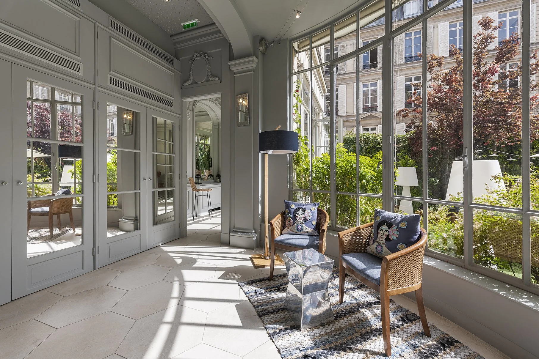 Salon lumineux avec grandes fenêtres, fauteuils en rotin avec coussins décorés représentant des femmes, tapis multicolore, lampe sur pied, et vue sur un jardin extérieur avec arbres et mobilier urbain, Cercle d'Aumale, hôtel particulier, paris, 