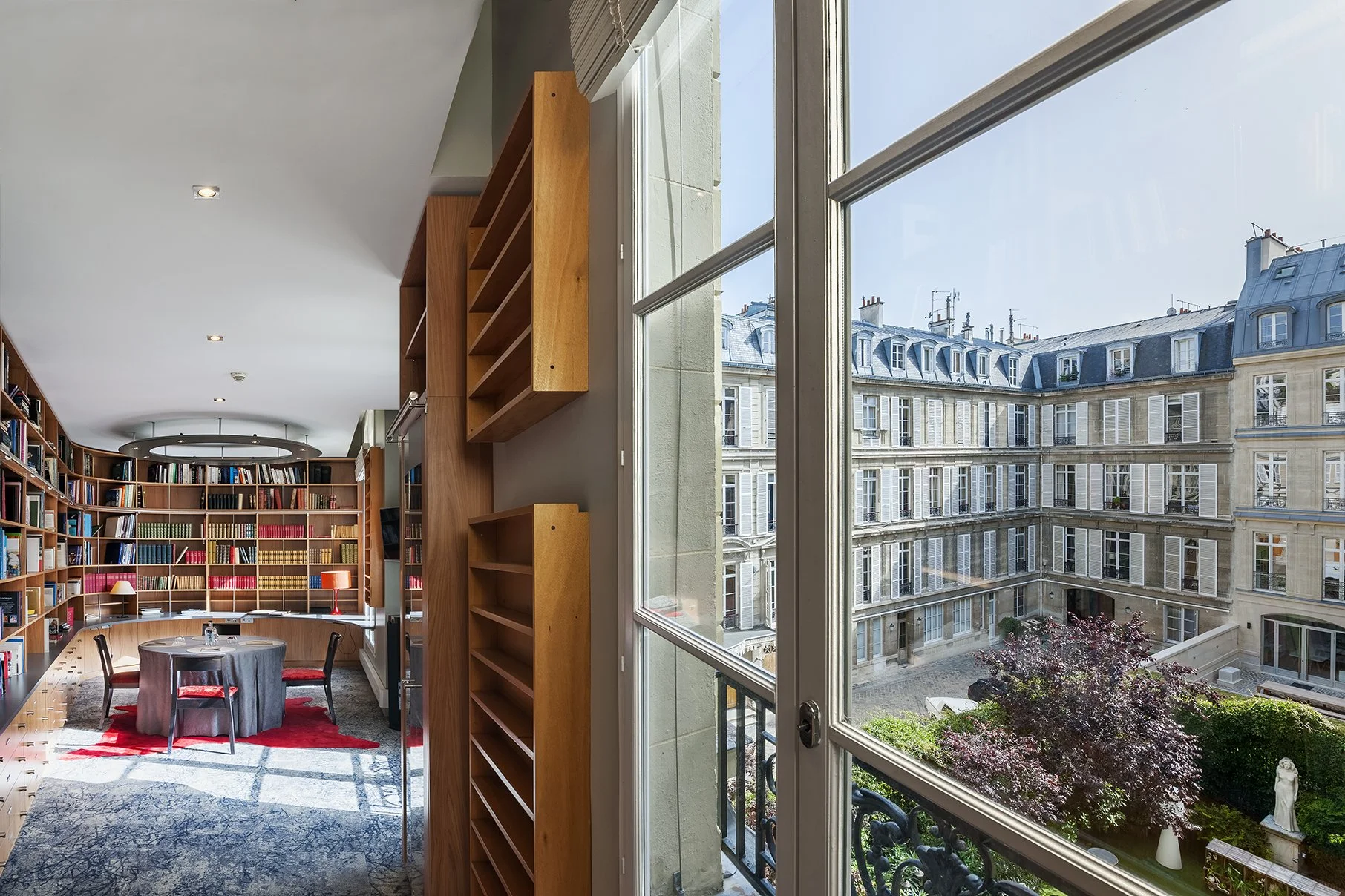 Une bibliothèque moderne avec murs en bois et grande fenêtre offrant une vue sur un immeuble parisien avec des fenêtres à volets bleus, un patio et des arbres, Cercle d'Aumale, hôtel particulier, Paris, Location, Prestige, Luxe, Dalida,