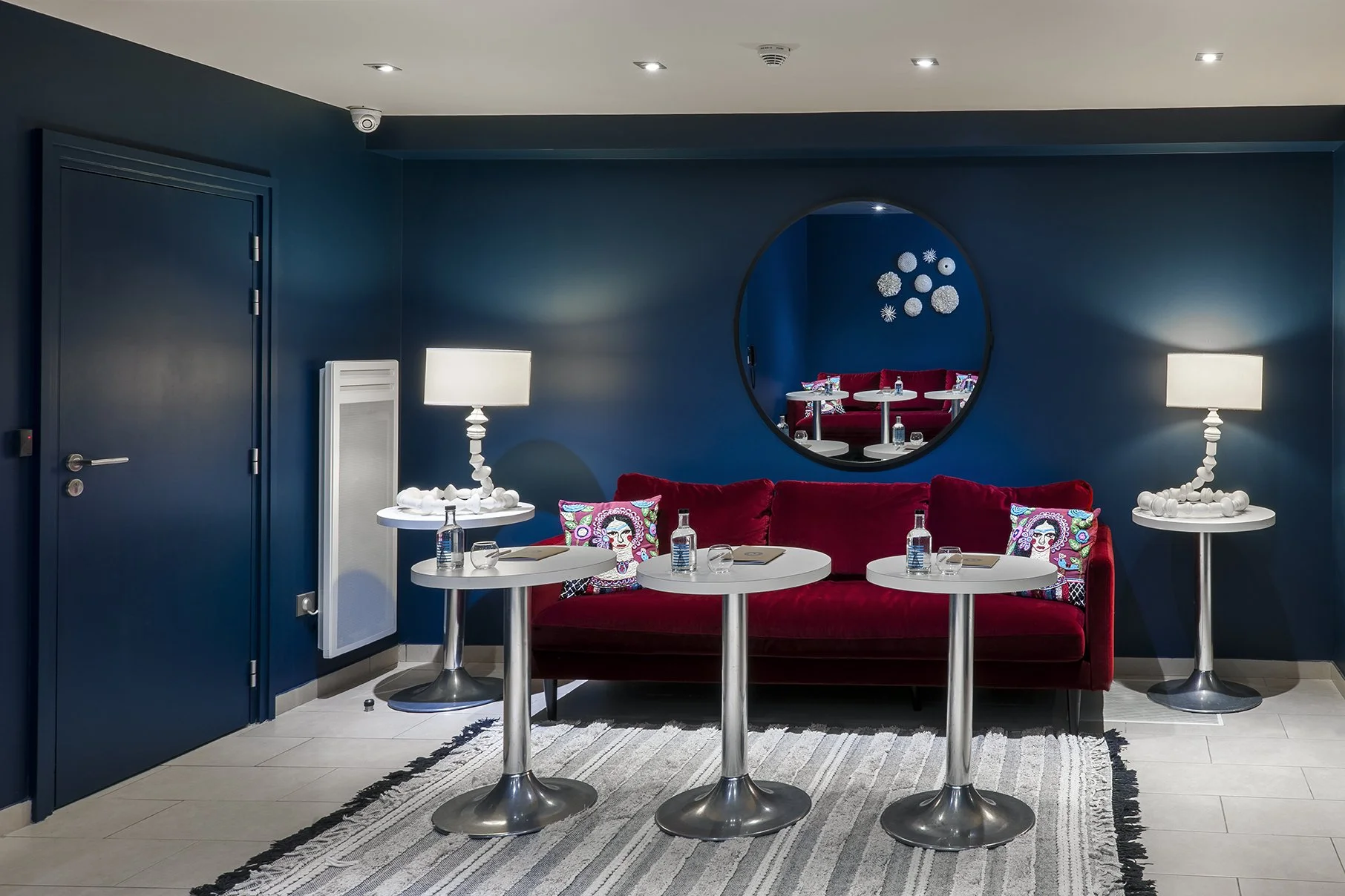 Une salle de réception avec un canapé rouge, deux lampes blanches, plusieurs tables rondes, et des coussins colorés au mur bleu, avec un miroir rond, Cercle d'Aumale, hôtel particulier, Paris, Location, Prestige, Luxe, Dalida,