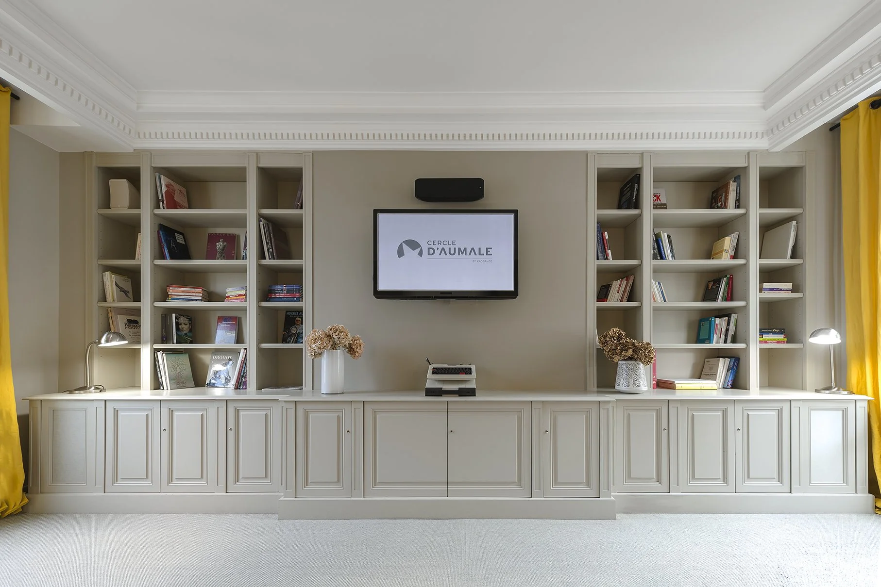 Une bibliothèque blanche avec des étagères remplies de livres, deux lampes de bureau, deux vases avec des fleurs séchées, un écran plat avec le logo cercle d'AUMLE, Cercle d'Aumale, hôtel particulier, Paris, Location, Prestige, Luxe, Dalida,