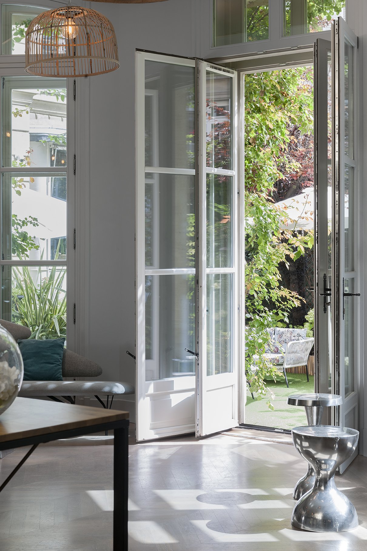 Intérieur avec une porte-fenêtre s'ouvrant sur un jardin, un canapé avec un coussin bleu, une lampe en rotin suspendue au plafond, une table en verre avec une sculpture en acier inoxydable, Cercle d'Aumale, hôtel particulier, Luxe, Dalida, Paris,