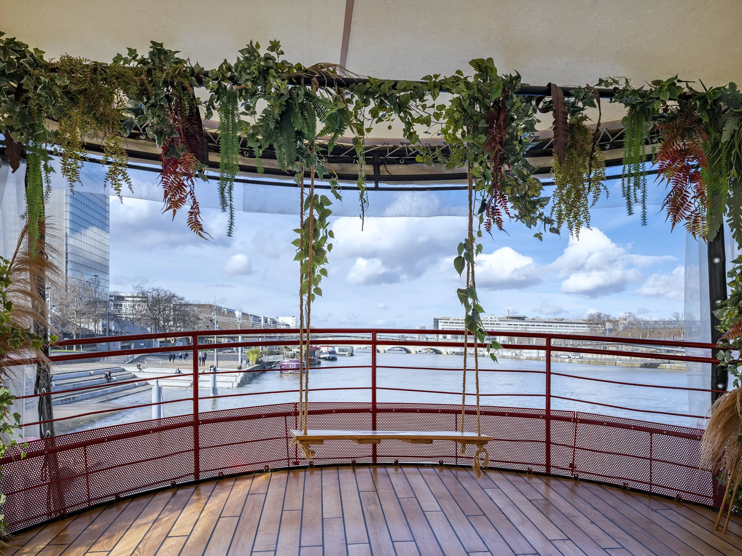 Une vue depuis un bateau avec une balancelle en bois suspendue, entouré de plantes vertes suspendues, avec un fond de ciel bleu et des bâtiments modernes au loin, Bateau Phare, Paris, Concert, Nightlife, Space Invaders, Mosaïc, Bar, Food, terrasse, 