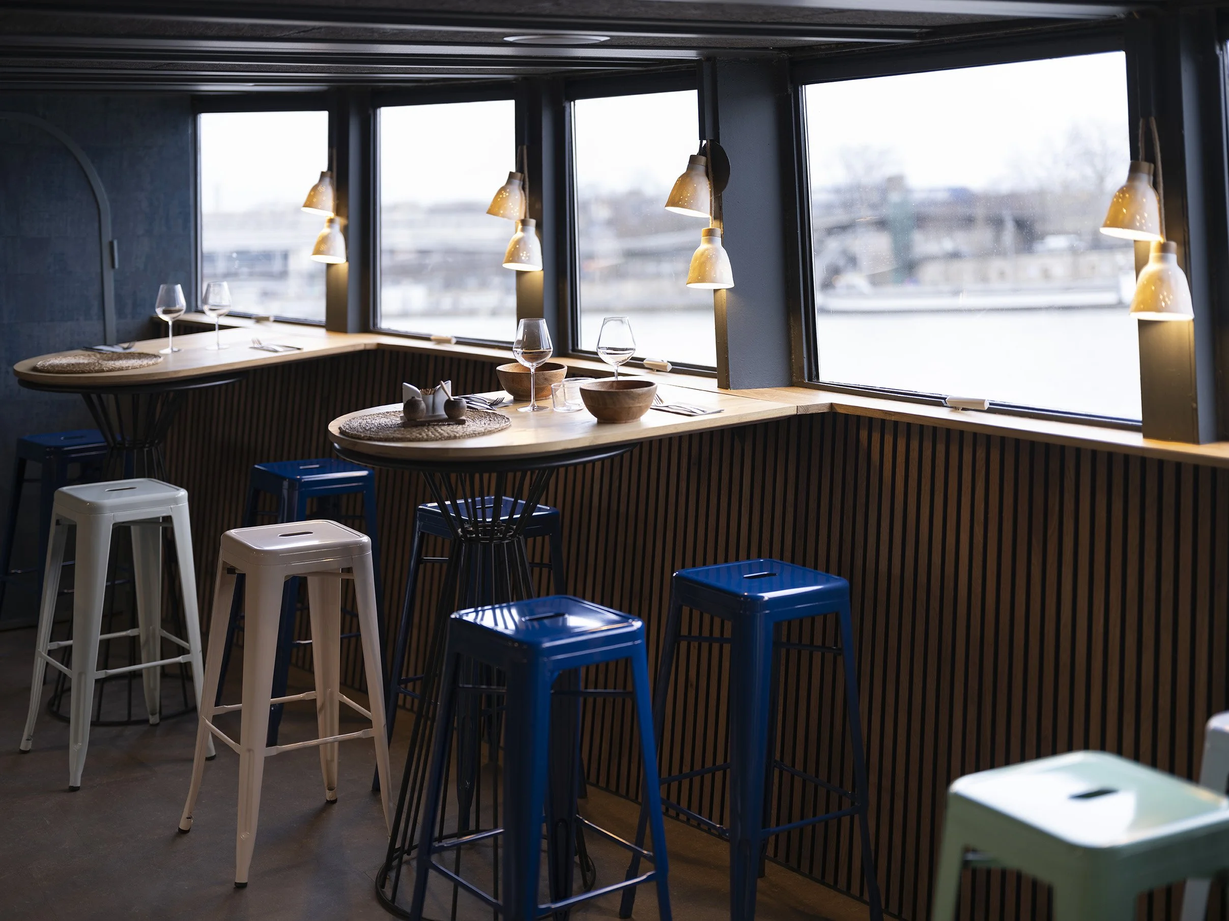 Intérieur d'un restaurant avec des tables, des chaises colorées, et des lampes suspendues au plafond, près de grandes fenêtres offrant une vue sur l'extérieur,  Bateau Phare, Paris, Concert, Nightlife, Space Invaders, Mosaïc, Bar, Food, terrasse, 