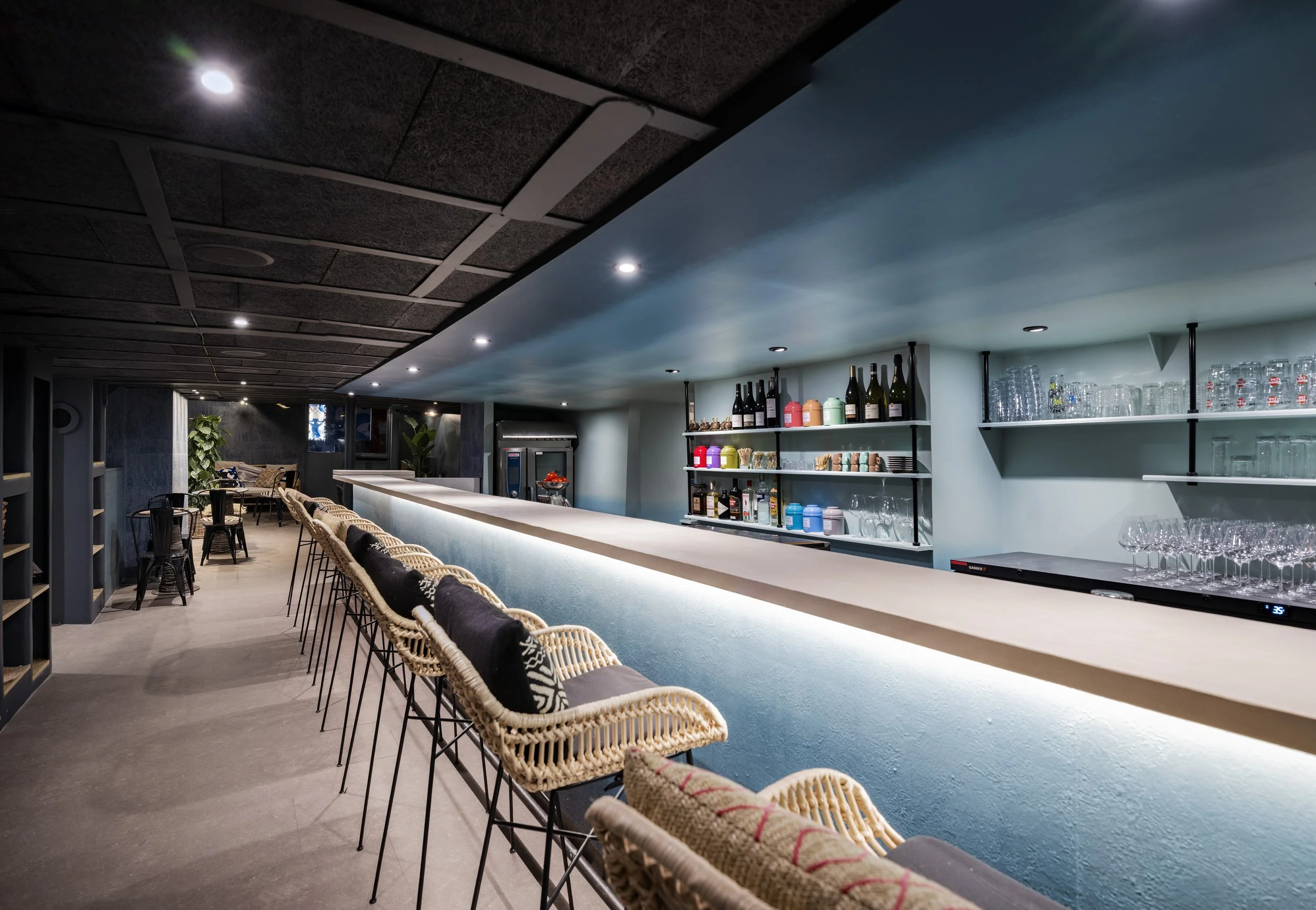 Intérieur d'un bar avec un comptoir, des chaises en rotin, étagères avec des bouteilles, des verres et des objets décoratifs, ambiance moderne et chaleureuse,   Bateau Phare, Paris, Concert, Nightlife, Mosaïc, Bar, Food, terrasse, quai de Seine