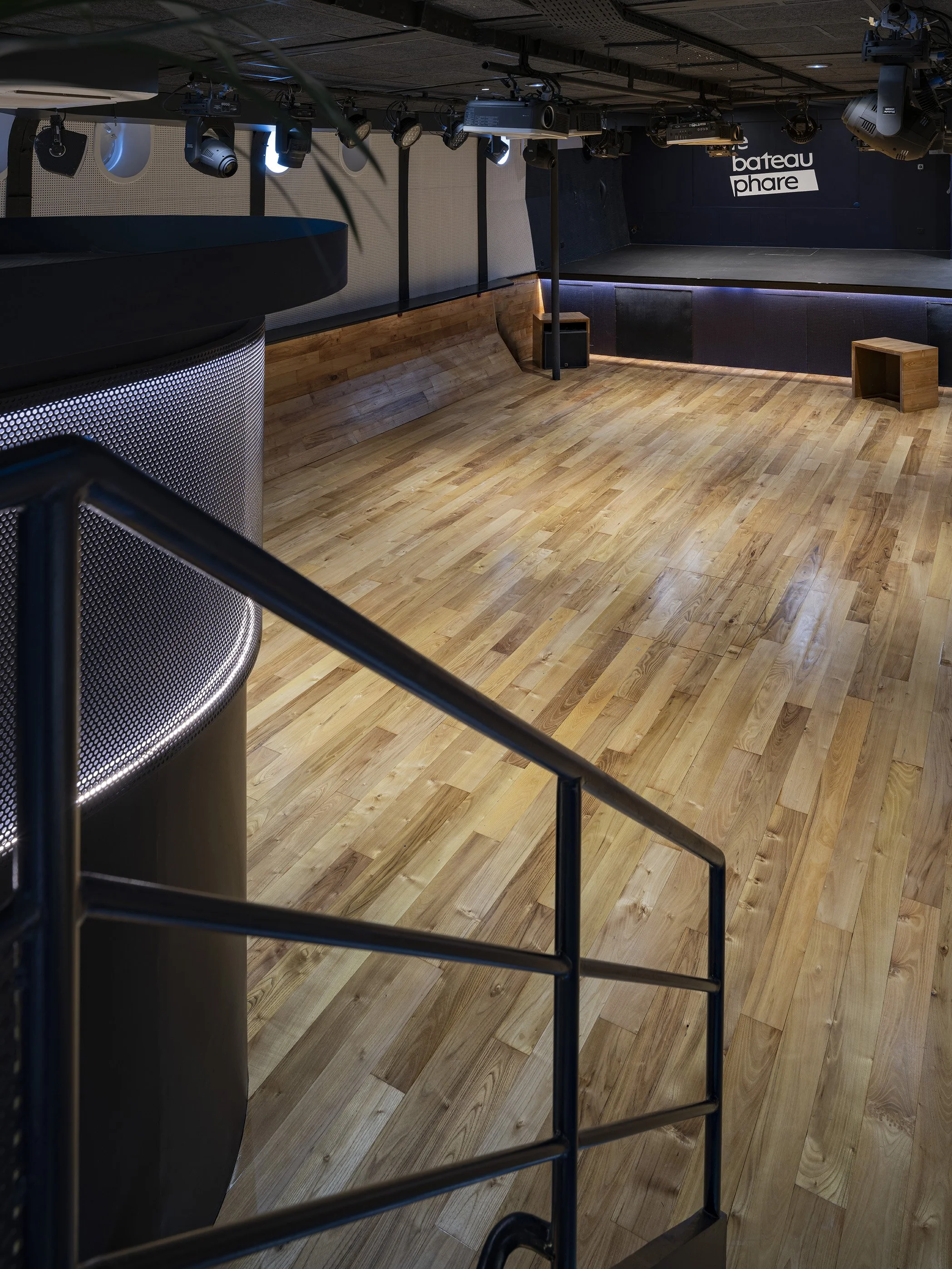 Salle de spectacle ou scène avec éclairage et équipements audio, avec un sol en bois clair et des murs sombres, et une bannière avec le texte 'bateau phare'.