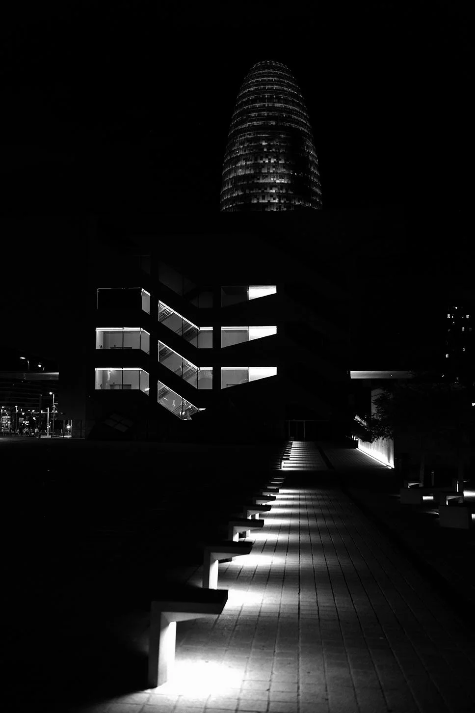 Une scène nocturne en noir et blanc avec un bâtiment moderne éclairé, un chemin bordé de lampes, et une tour en arrière-plan. architecture, musée du design, Barcelone, Espagne, nuit,