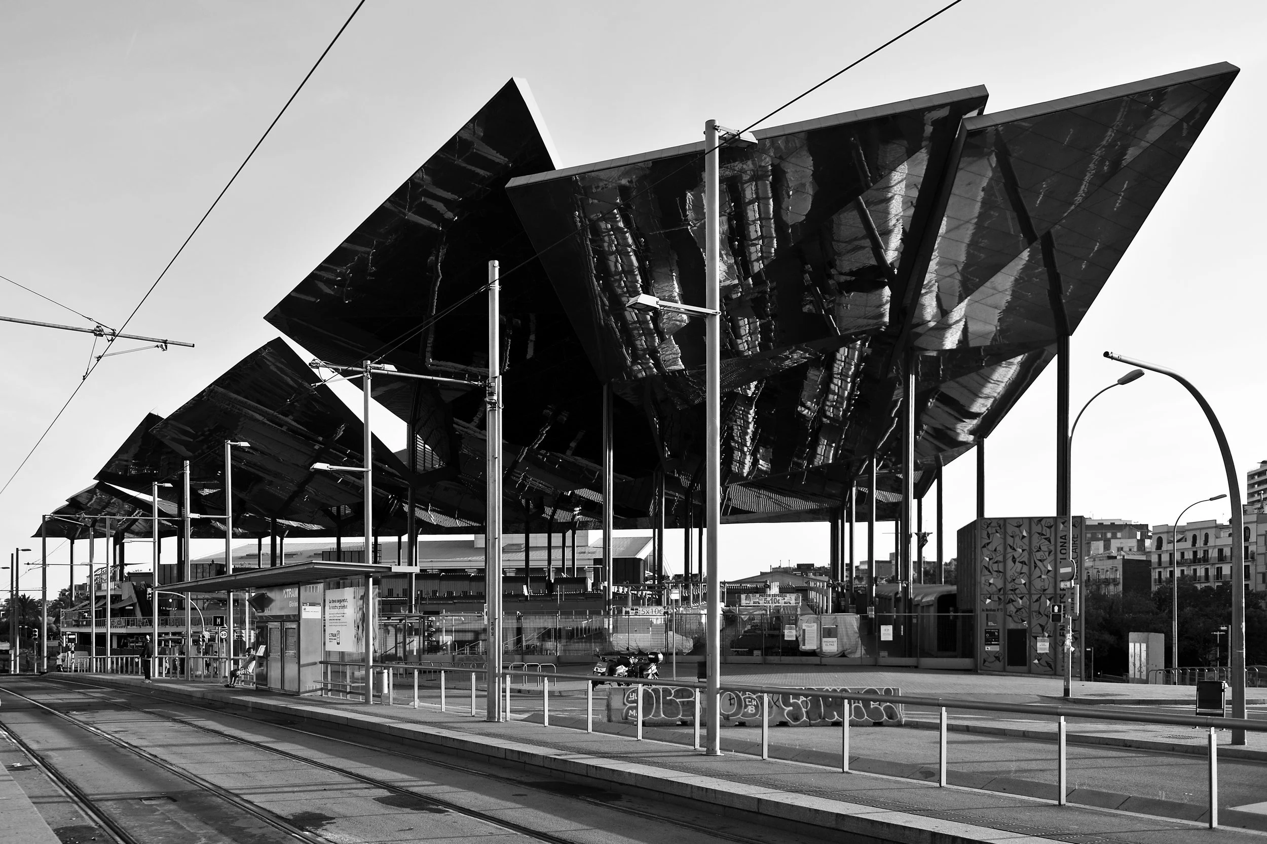Une station de tramway moderne avec une structure en acier et en verre, des lignes de tramway, des poteaux électriques, des lampadaires et des bâtiments en arrière-plan, en noir et blanc. Barcelone, Spain, 