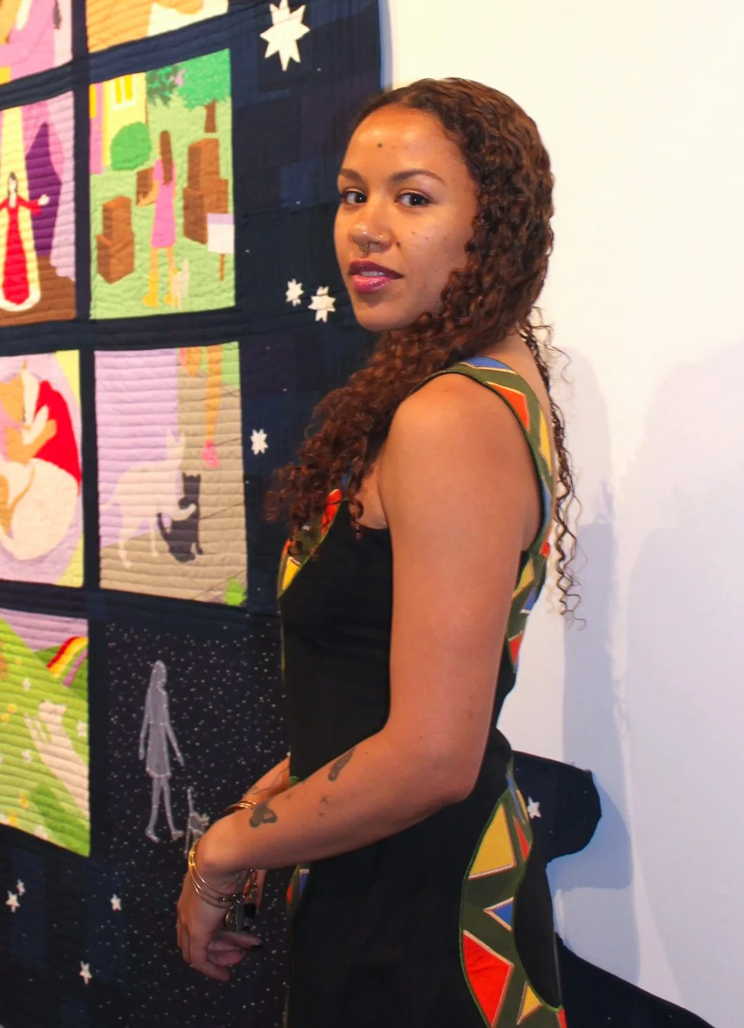 A mixed race young woman with long brown curly hair wearing a sleeveless black dress with geometric patterns softly smiles in front of a portion of a quilt. The quilt has illustrative squares surrounded by a navy border.