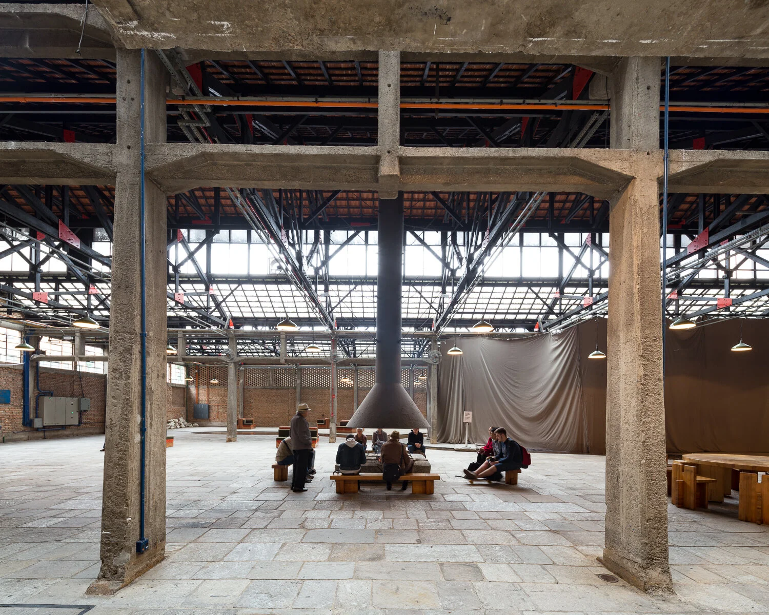 Interior of an industrial-style warehouse with exposed beams and large windows, featuring people sitting on benches around a central cone-shaped structure.