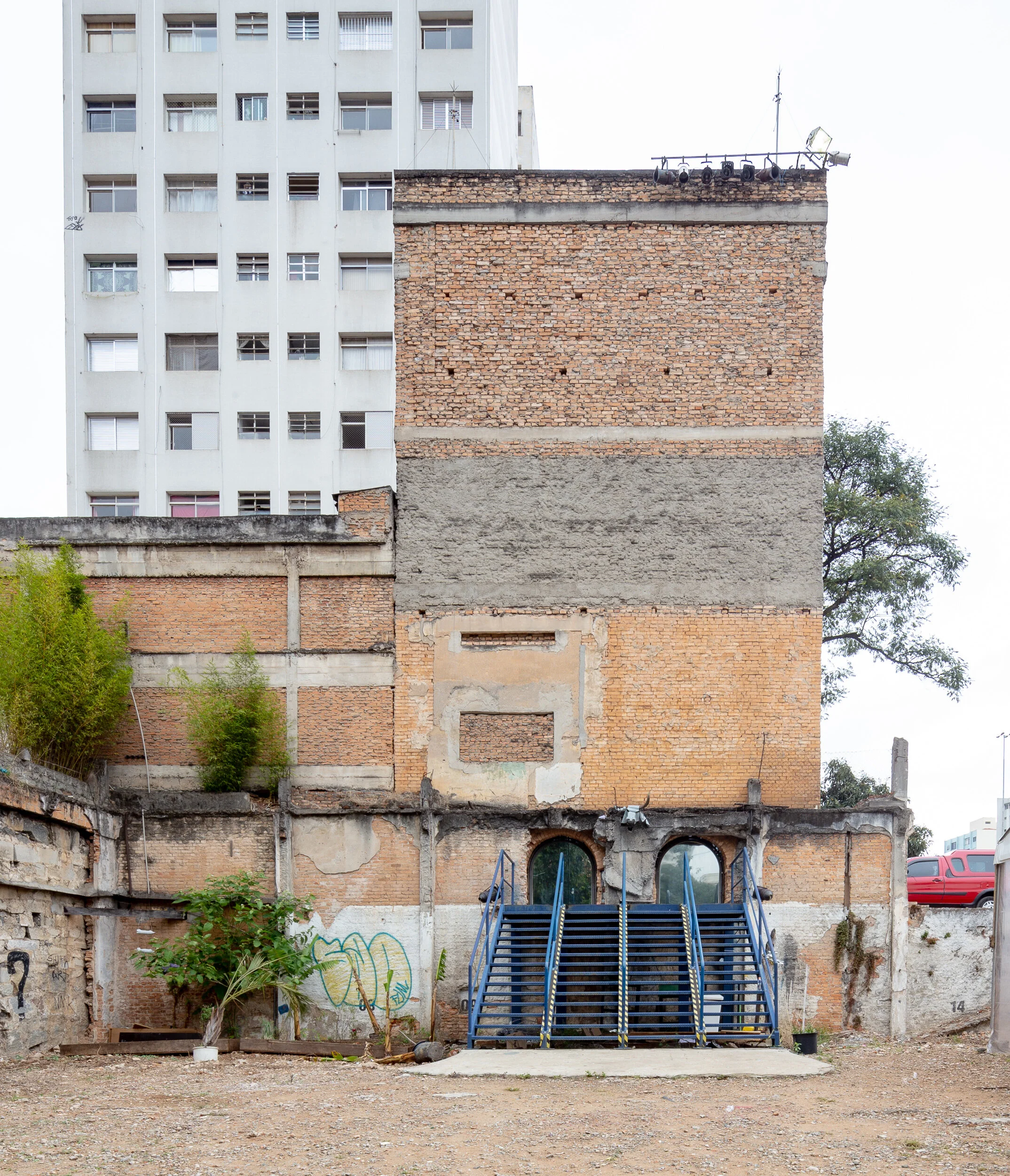 An old brick wall with a set of metal stairs in front, surrounded by sparse vegetation and a tall residential building in the background.