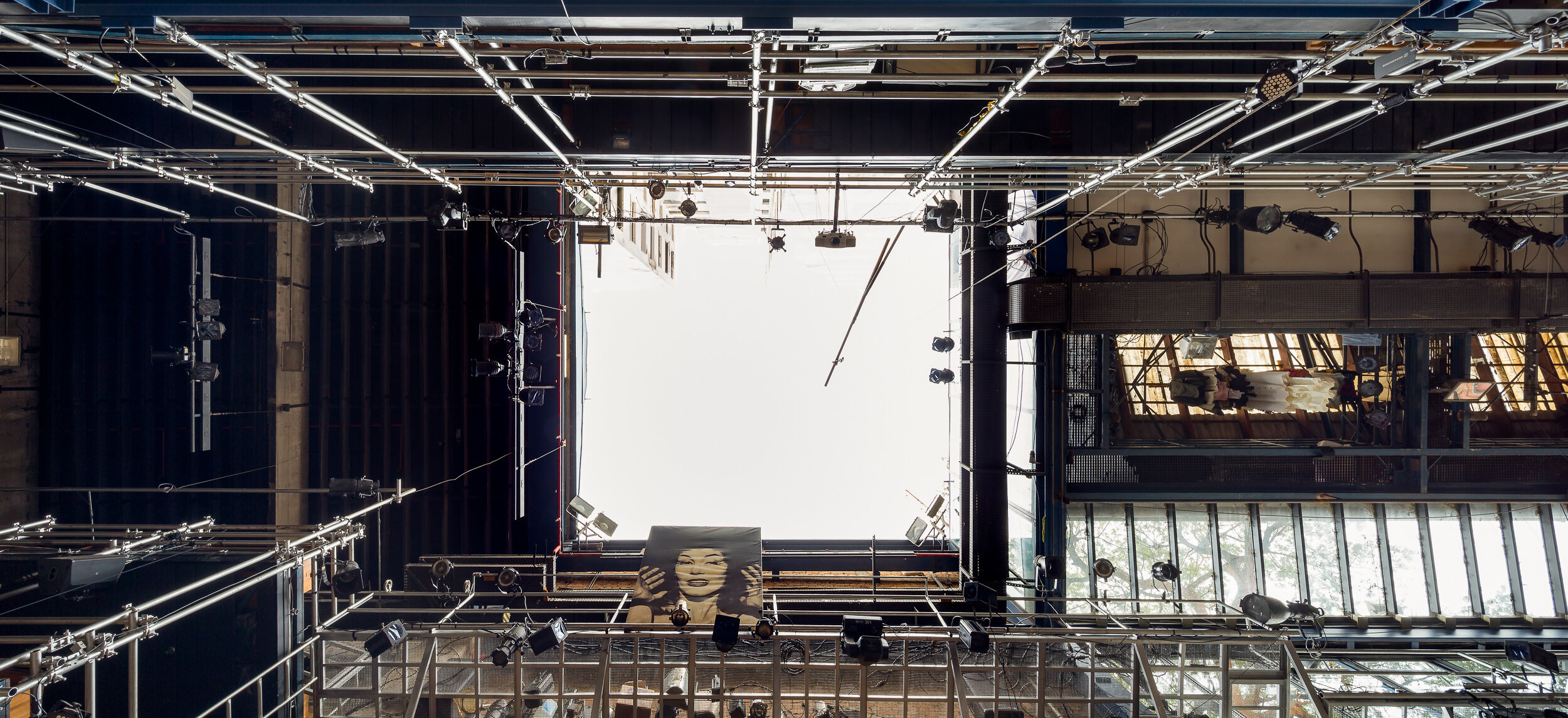 View from below of a theatrical stage rigging with lights and scaffolding, featuring a large portrait banner hanging in the center.