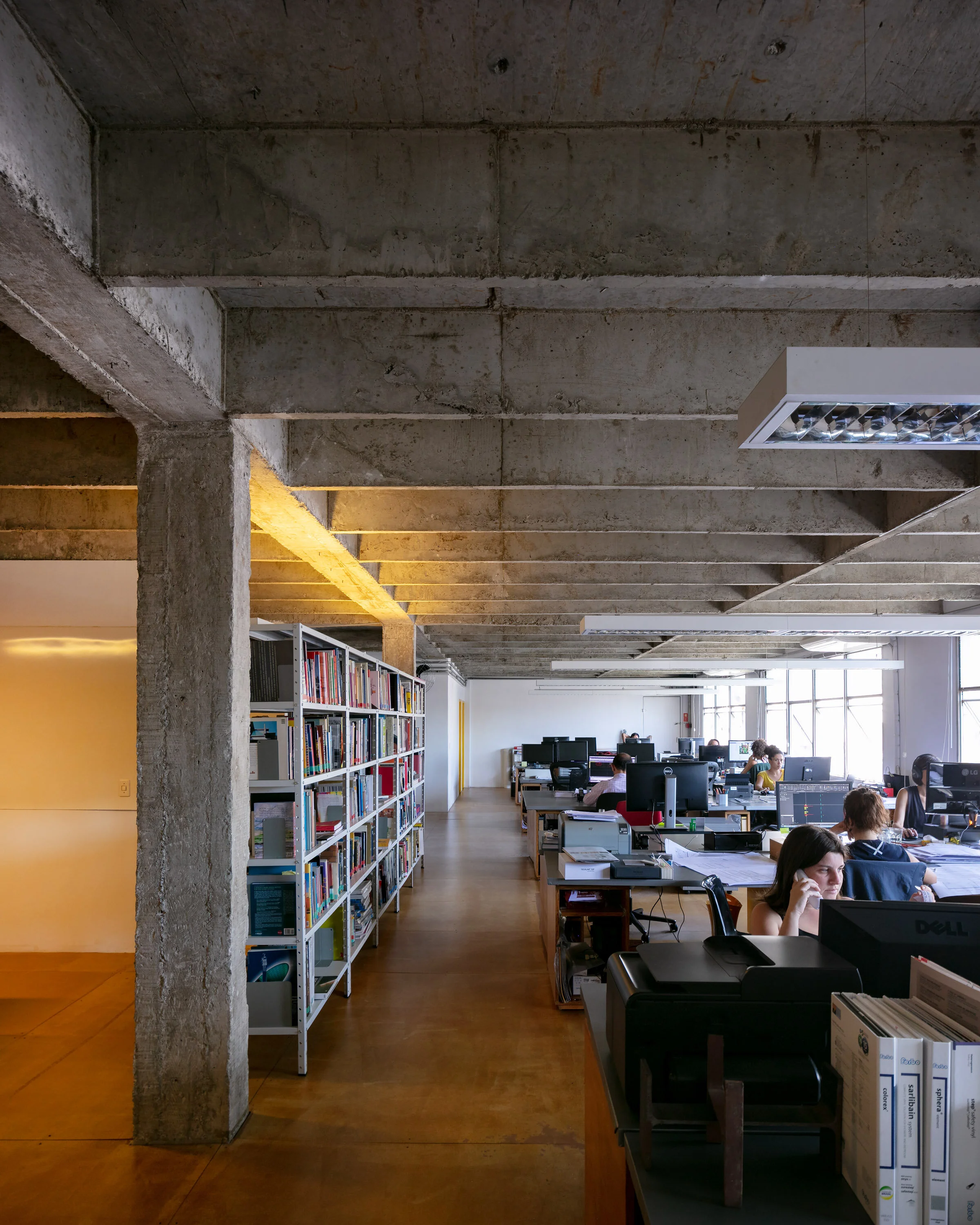Modern office space with open floor plan and exposed concrete ceiling, featuring desks with computers, bookshelves along the wall, and large windows providing natural light.