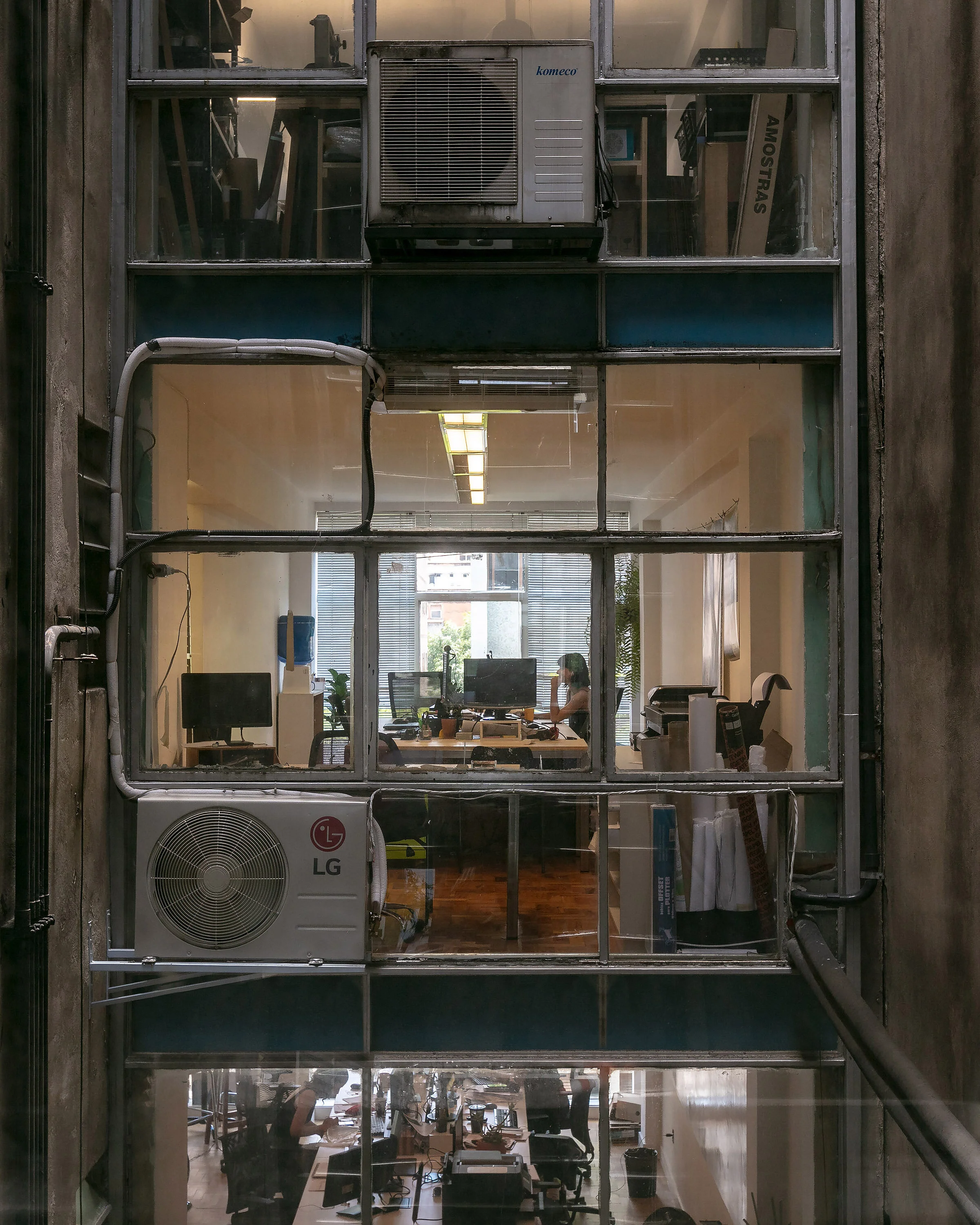 View through a window into an office space with multiple desks, computers, and people working inside. Air conditioning units are visible on the building exterior.