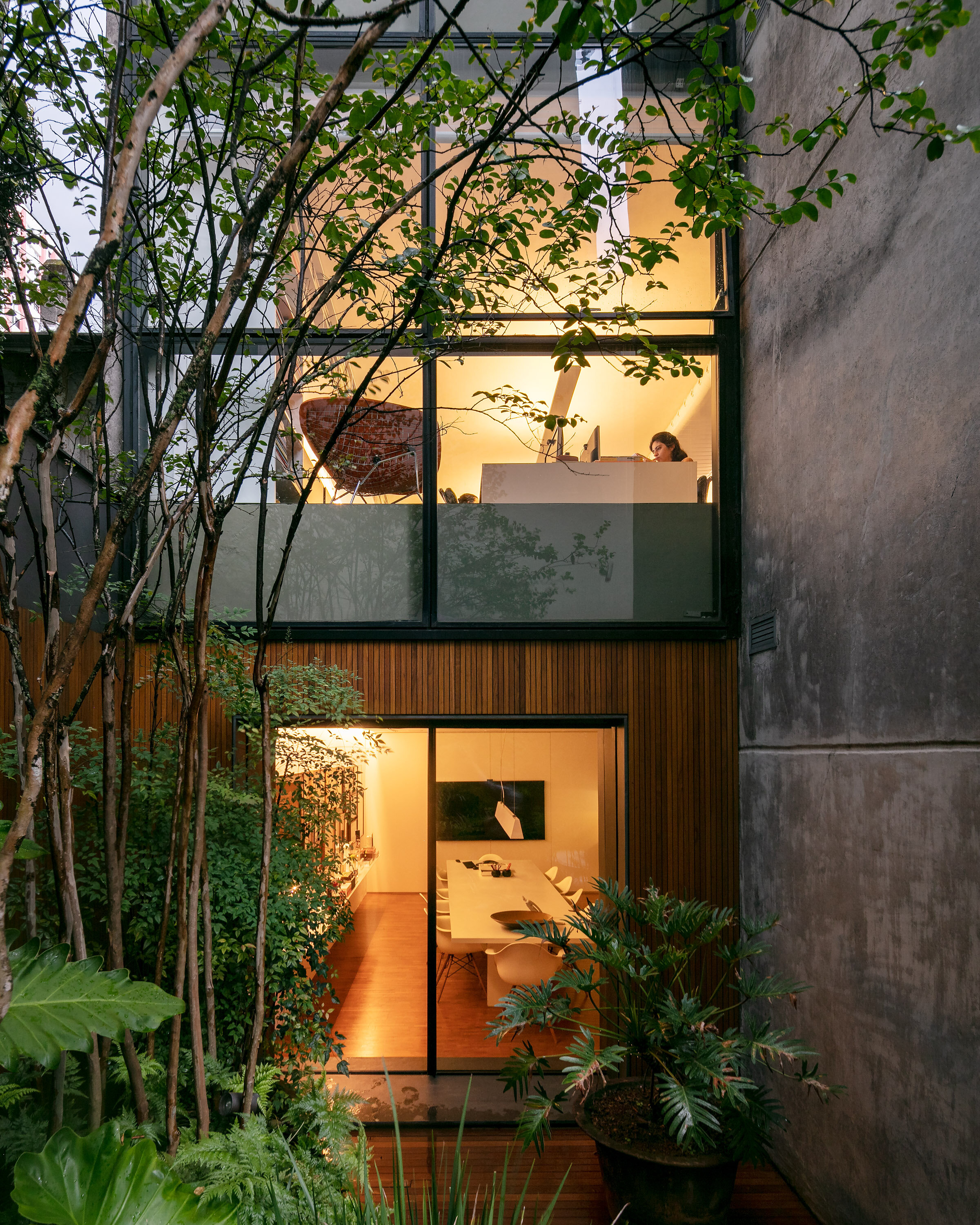 View of a modern two-story building with large glass windows. The bottom floor reveals a dining or conference room with a long table and chairs. The upper floor shows a workspace with a person working at a desk. The surrounding area has lush green pl