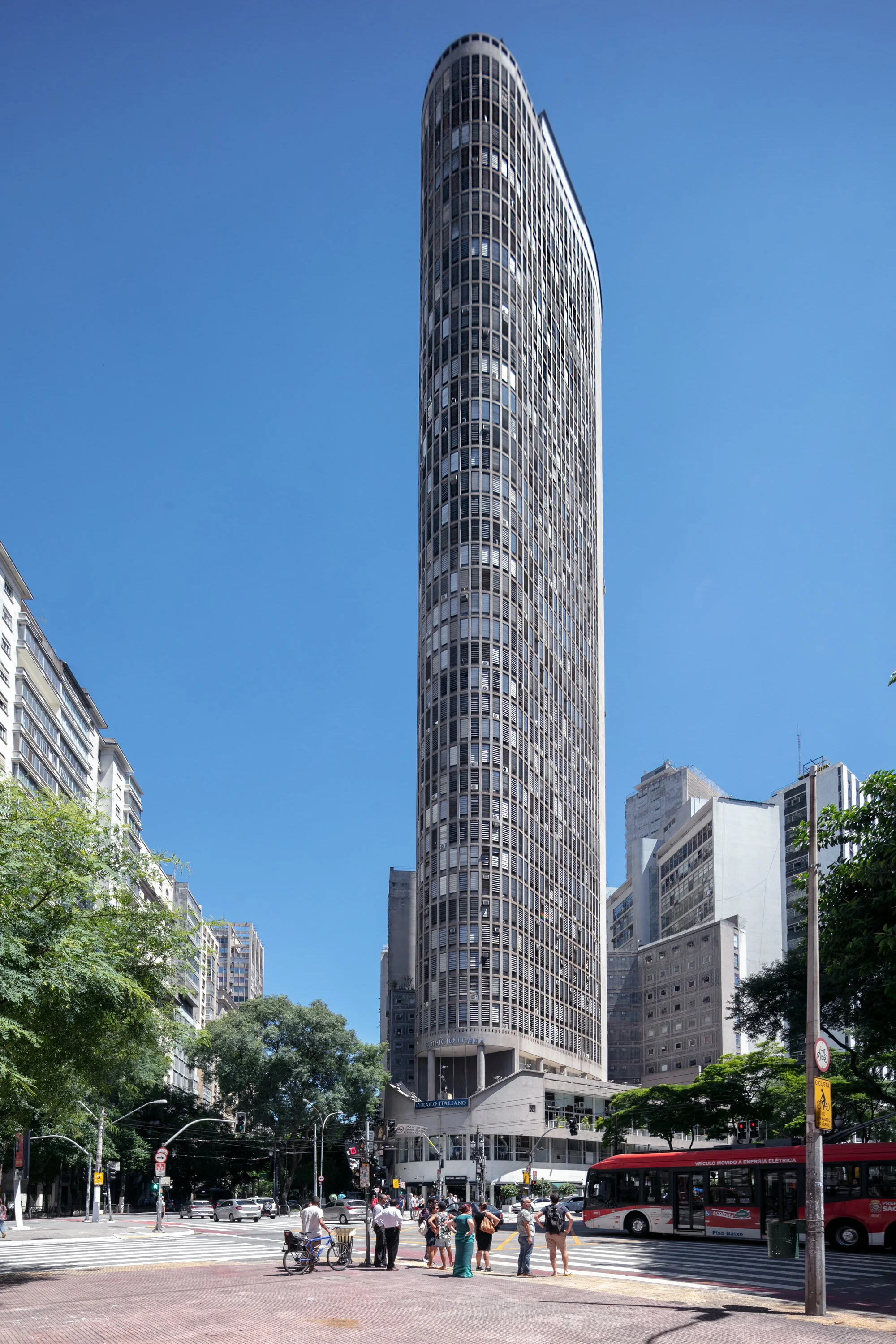A tall, cylindrical skyscraper with a modern architectural design in an urban setting, surrounded by other buildings, trees, and people at a crosswalk. A red bus and other vehicles are visible on the street below a clear, blue sky.