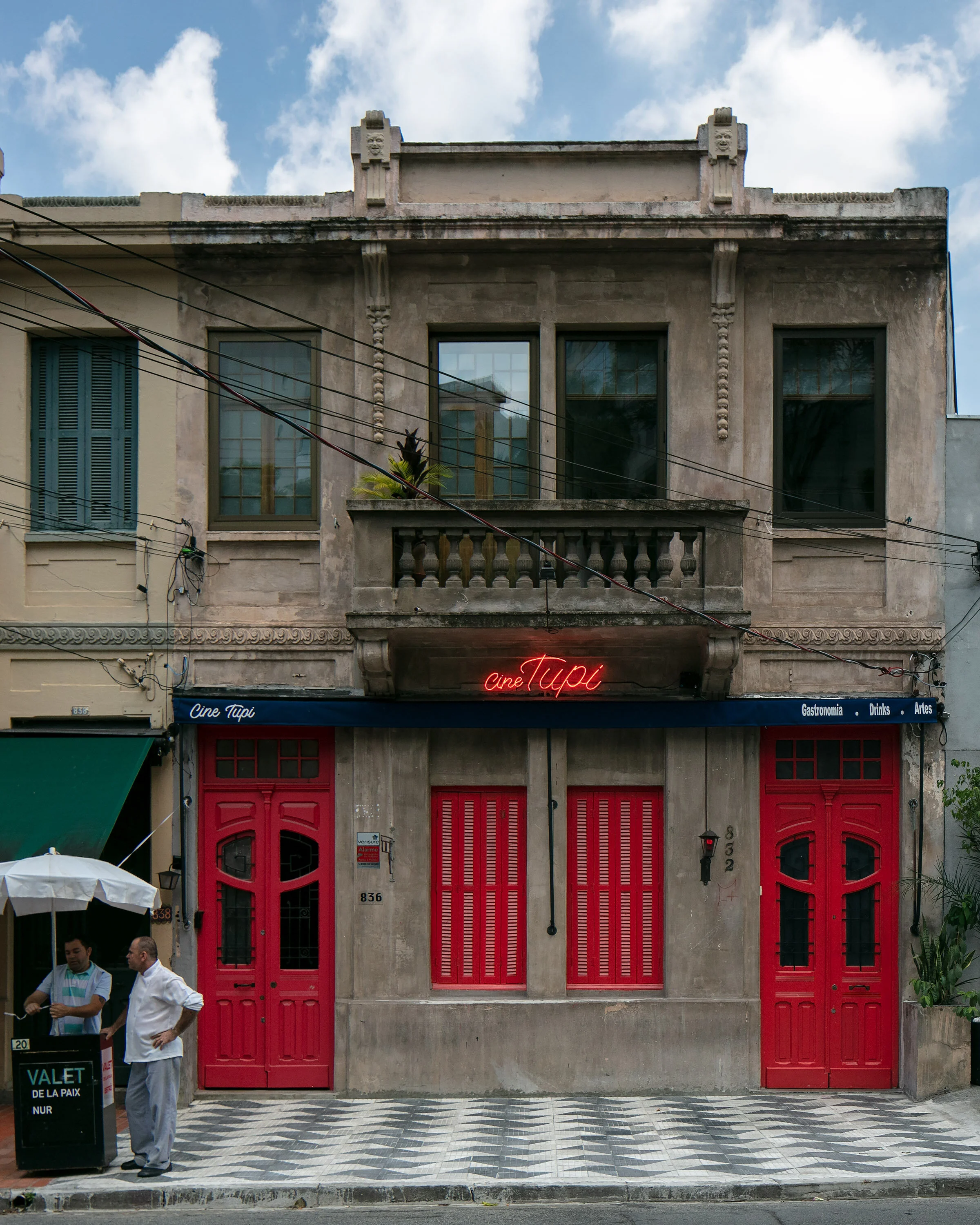 Historic building with red doors, neon sign "Cine Tupi," and patterned sidewalk, featuring two men talking under a street canopy.