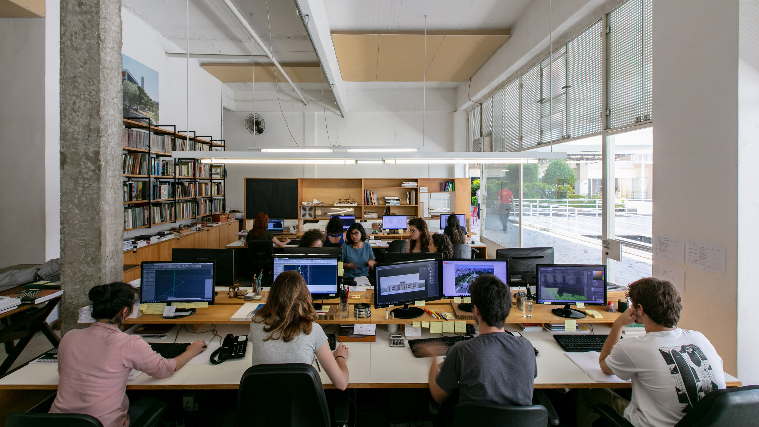 Open office space with multiple people working at computer desks, with shelves of books and large windows.