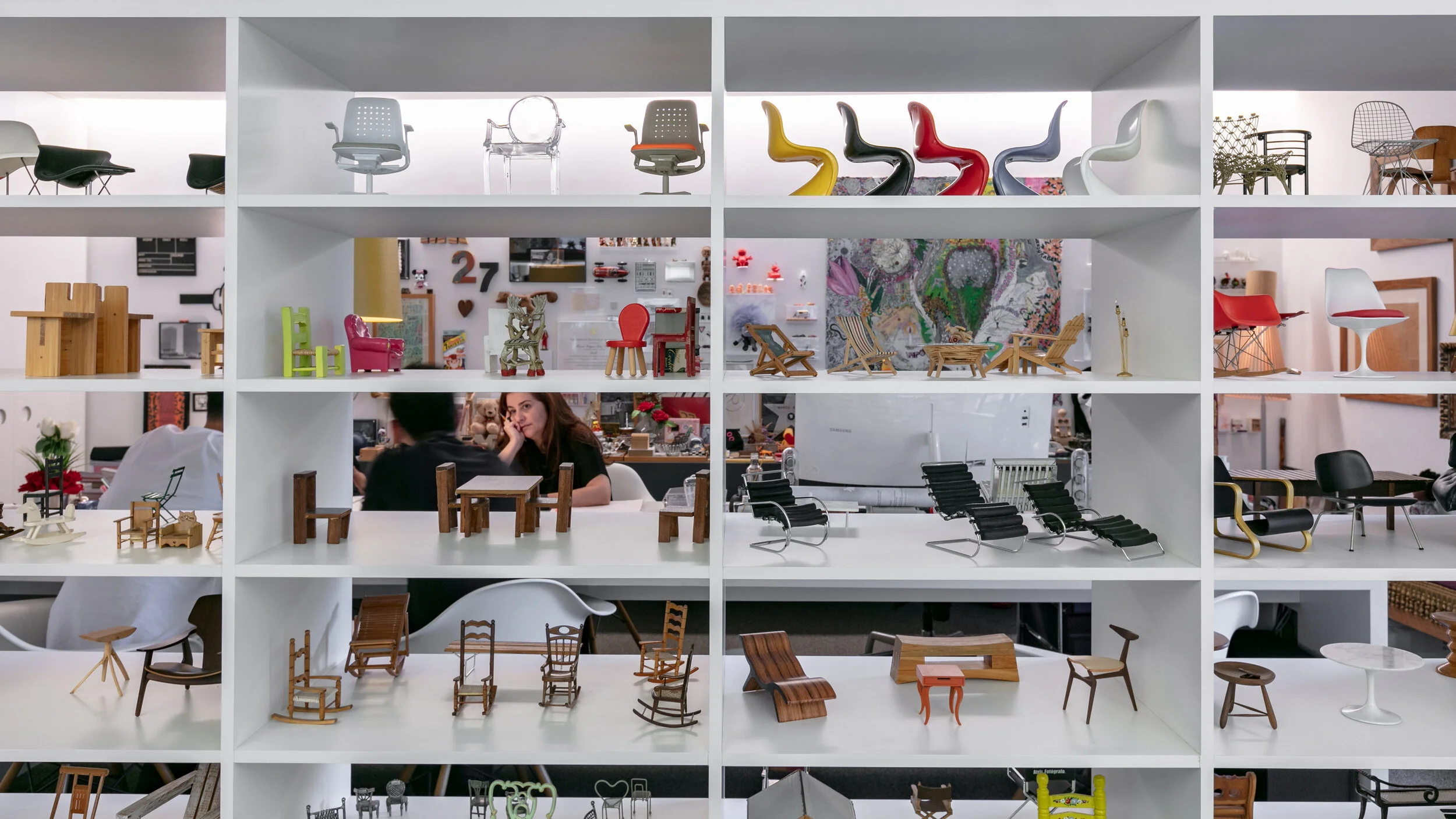 Wall shelves displaying miniature classic chairs and furniture models in a design studio with people working in the background.