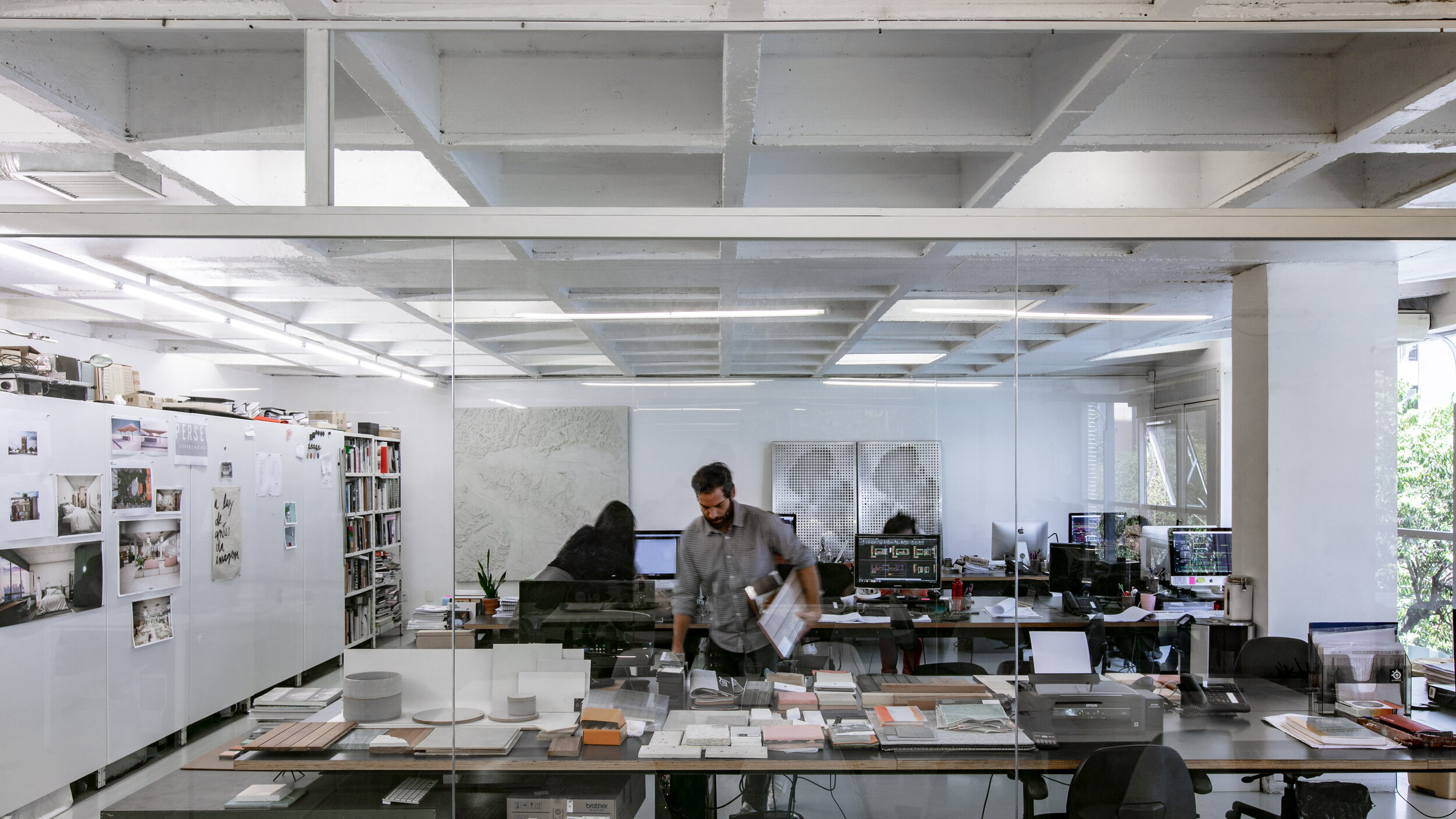 Interior of a modern office with a glass wall, showing several people working at desks with computers, architectural drawings, and design materials.