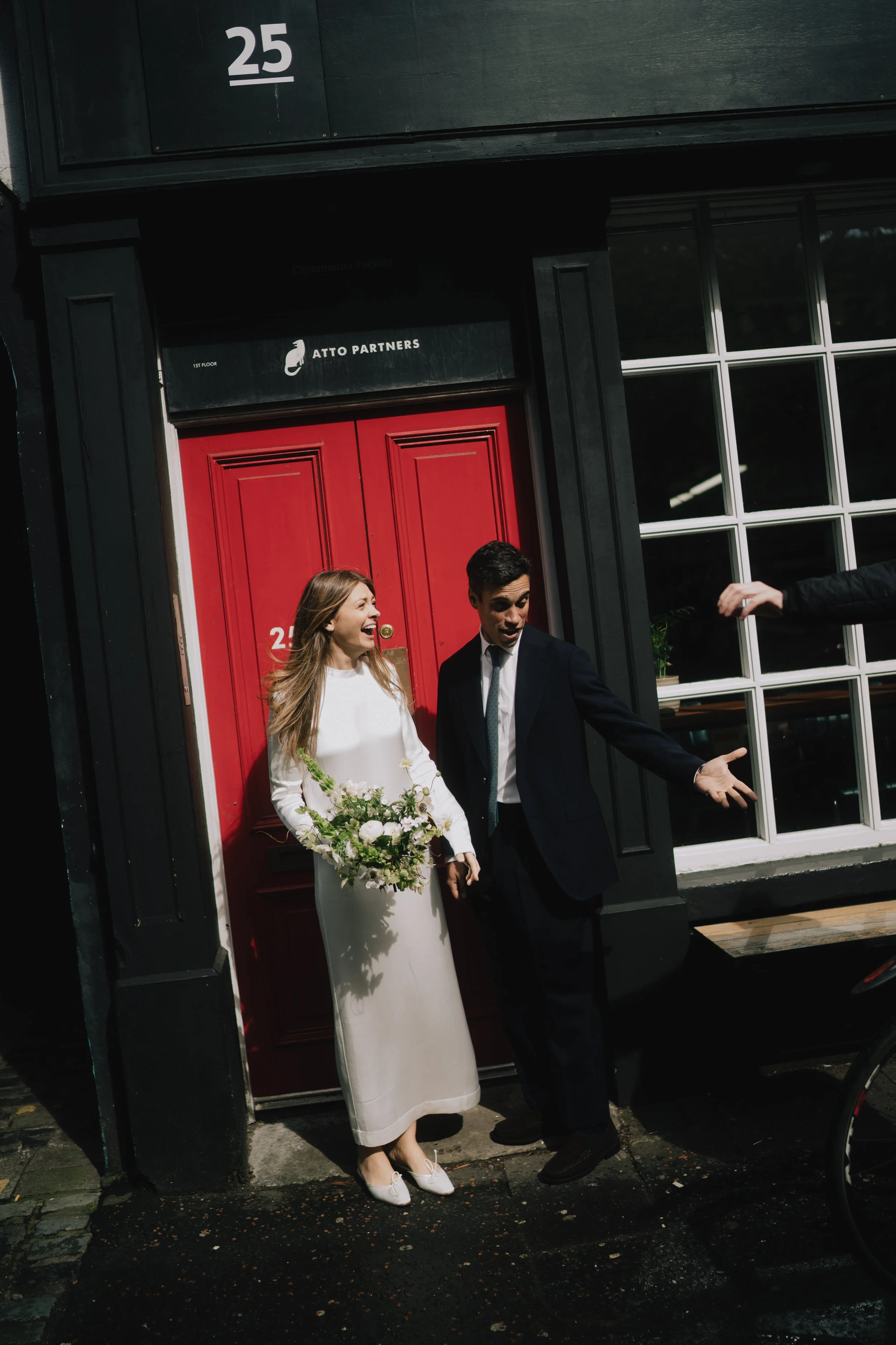 A newly married couple, a woman in a white dress holding a bouquet of flowers and a man in a suit, standing outside a black building with a red door, smiling and gesturing enthusiastically.