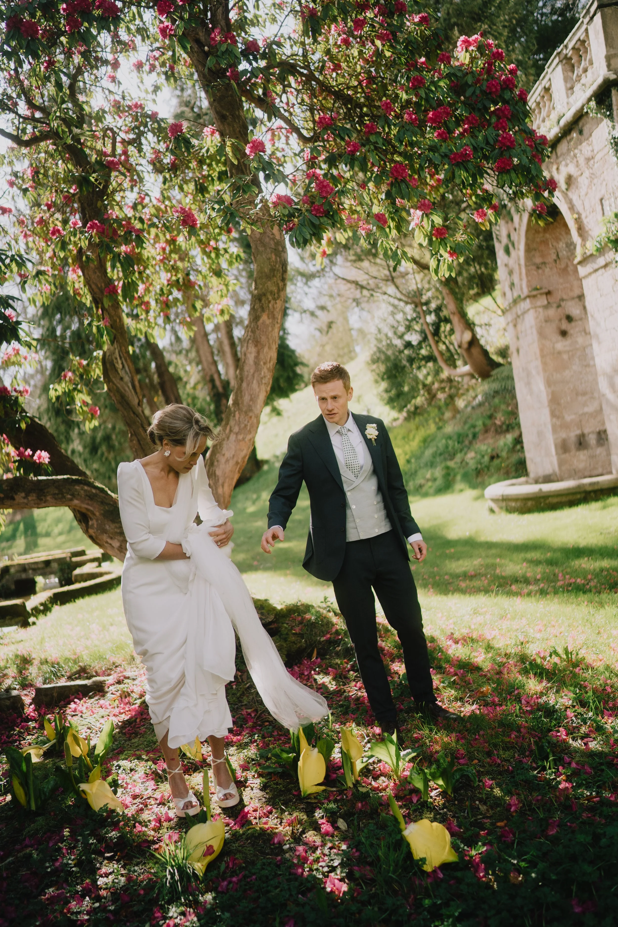 A bride in a white wedding dress and a groom in a black suit walk through a garden filled with pink flower petals, yellow calla lilies, and greenery, with a stone building in the background.