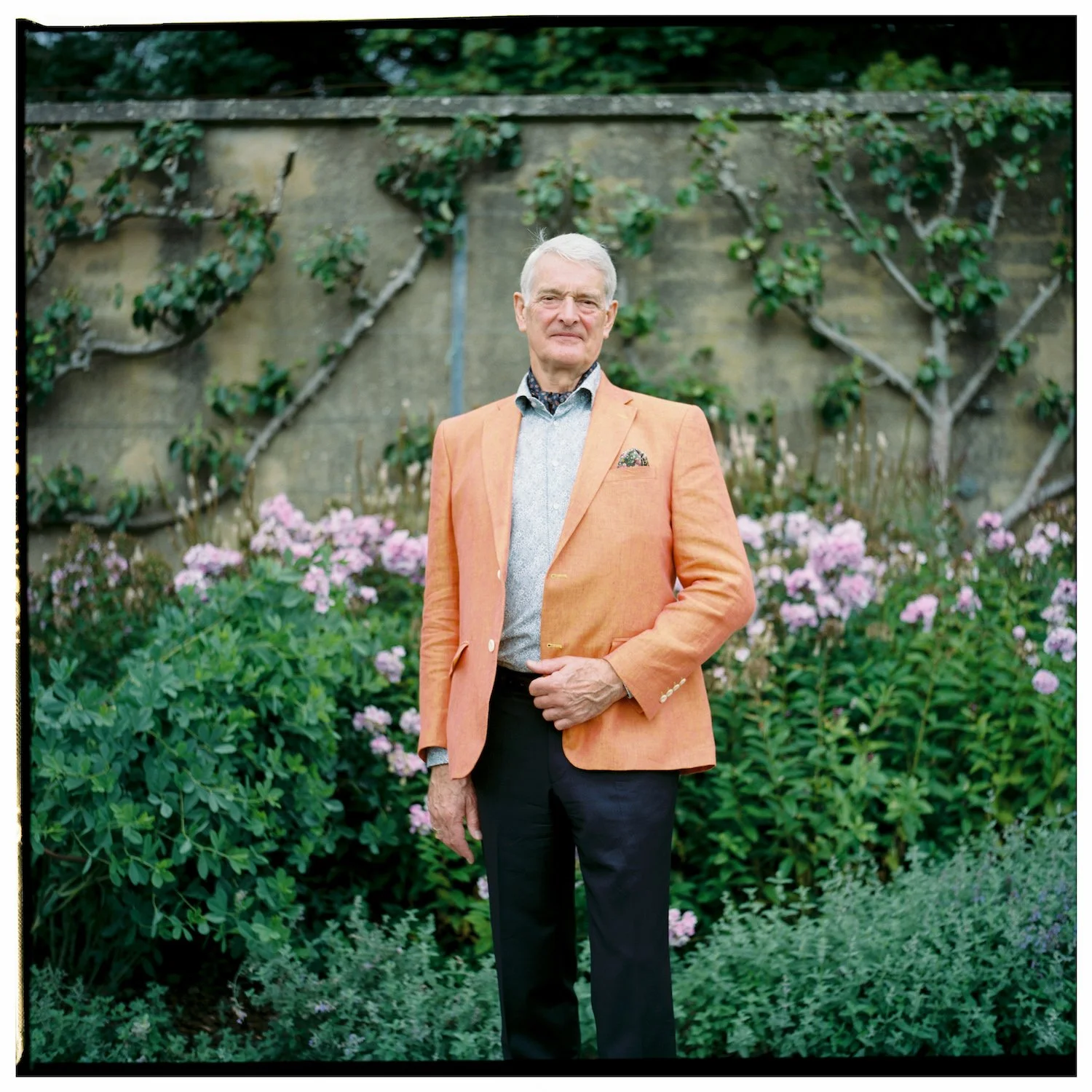 An elderly man standing in a garden with pink flowers and green foliage, wearing an orange blazer, light patterned shirt, and dark pants.