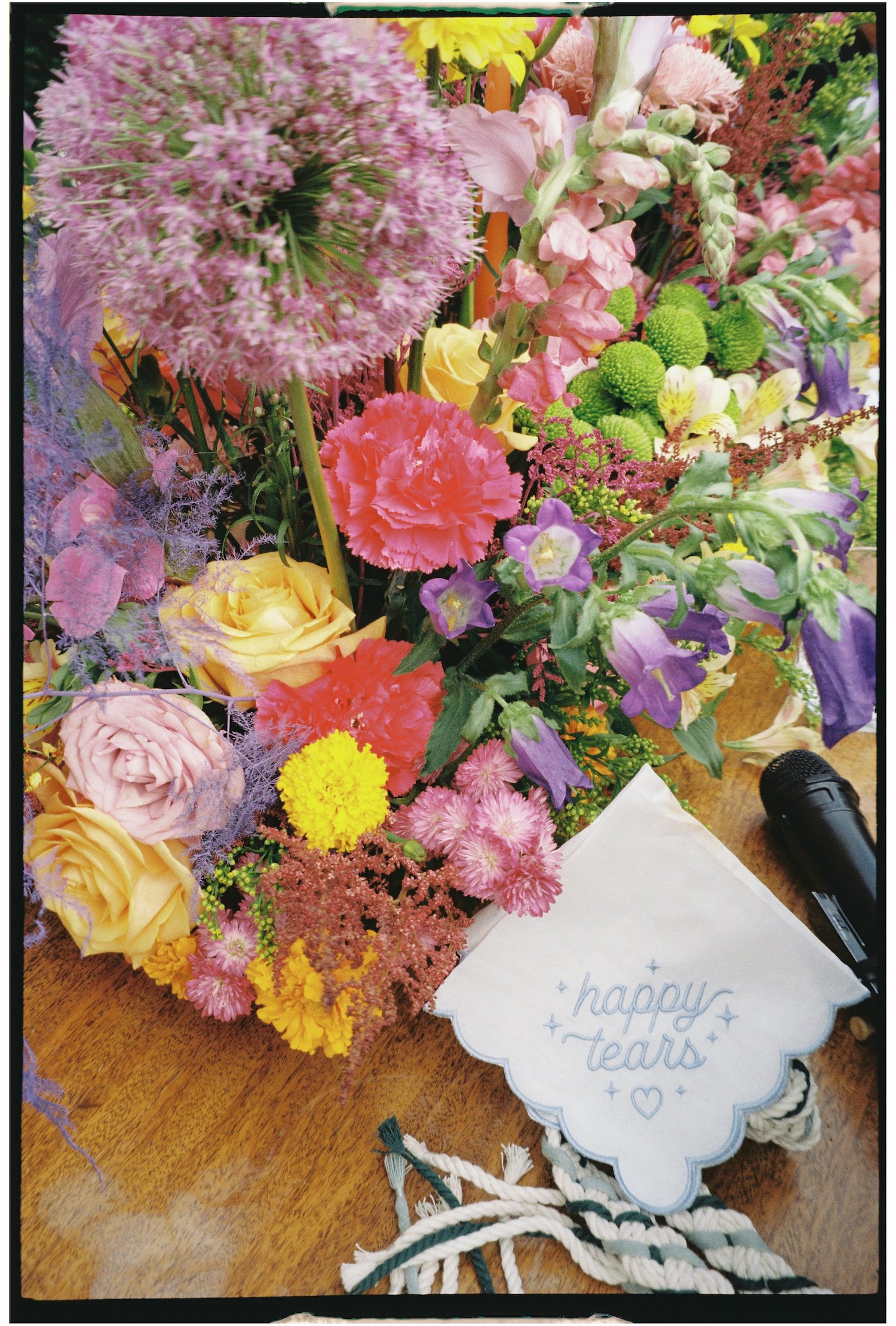 A large bouquet of colorful flowers on a wooden table, with a white paper decoration that says 'happy tears' and a black marker pen nearby.