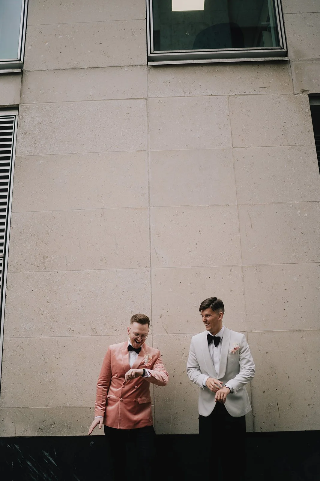 Two men dressed in tuxedos, one in a peach jacket and the other in a white jacket, standing against a beige stone wall, smiling and looking at each other.